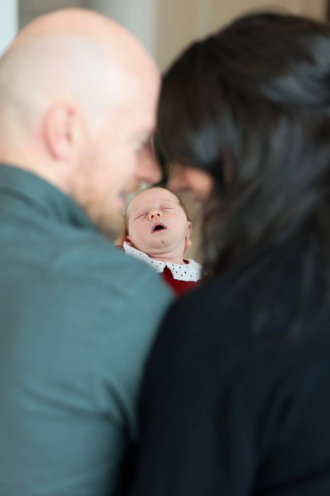 A baby being held up between a man and woman, with their foreheads touching and the baby sleeping with mouth open.