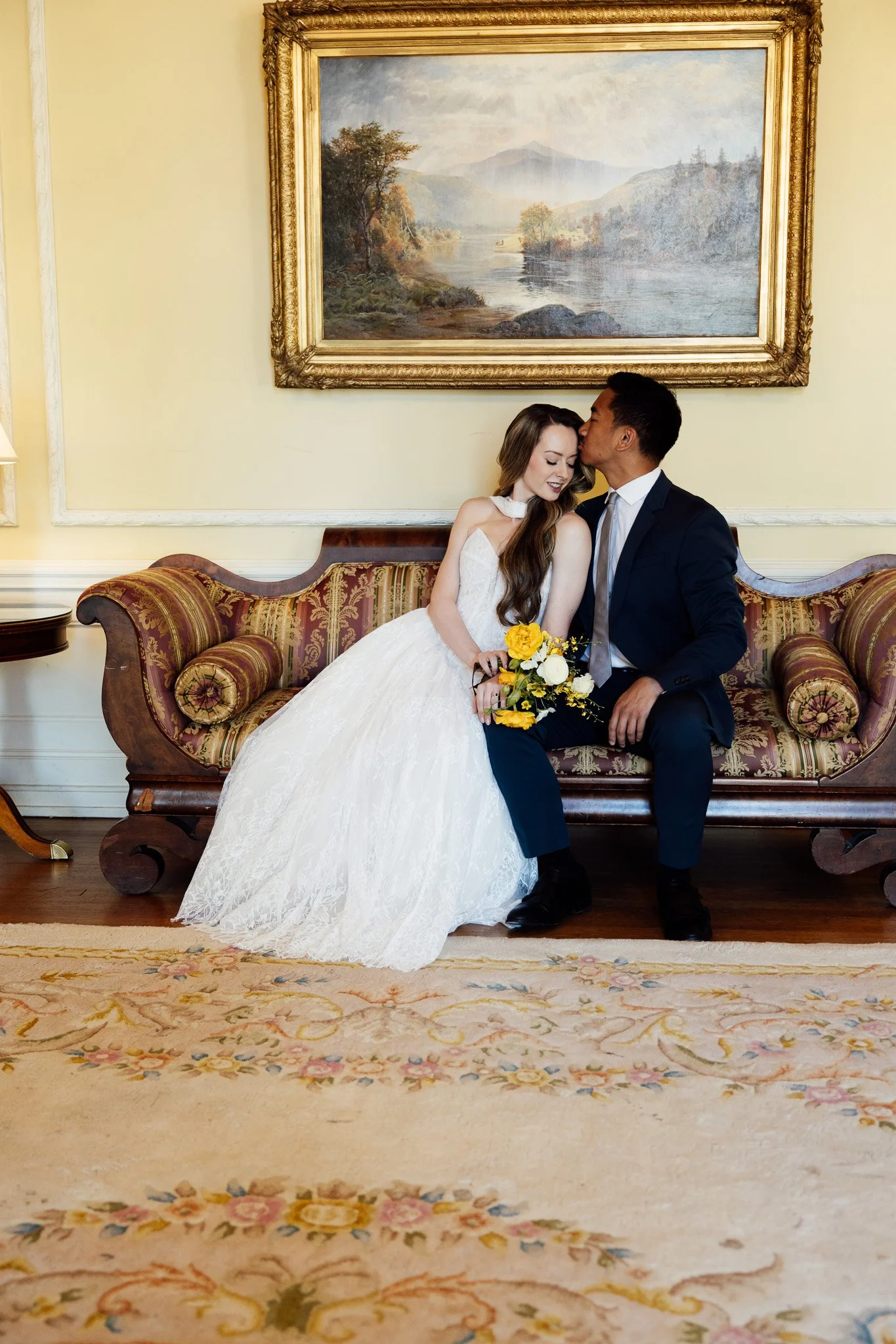 A couple dressed in wedding attire sitting on a vintage sofa in a room with a large framed landscape painting on the wall behind them. The woman is wearing a white wedding dress and holding a yellow bouquet, while the man is in a dark suit.