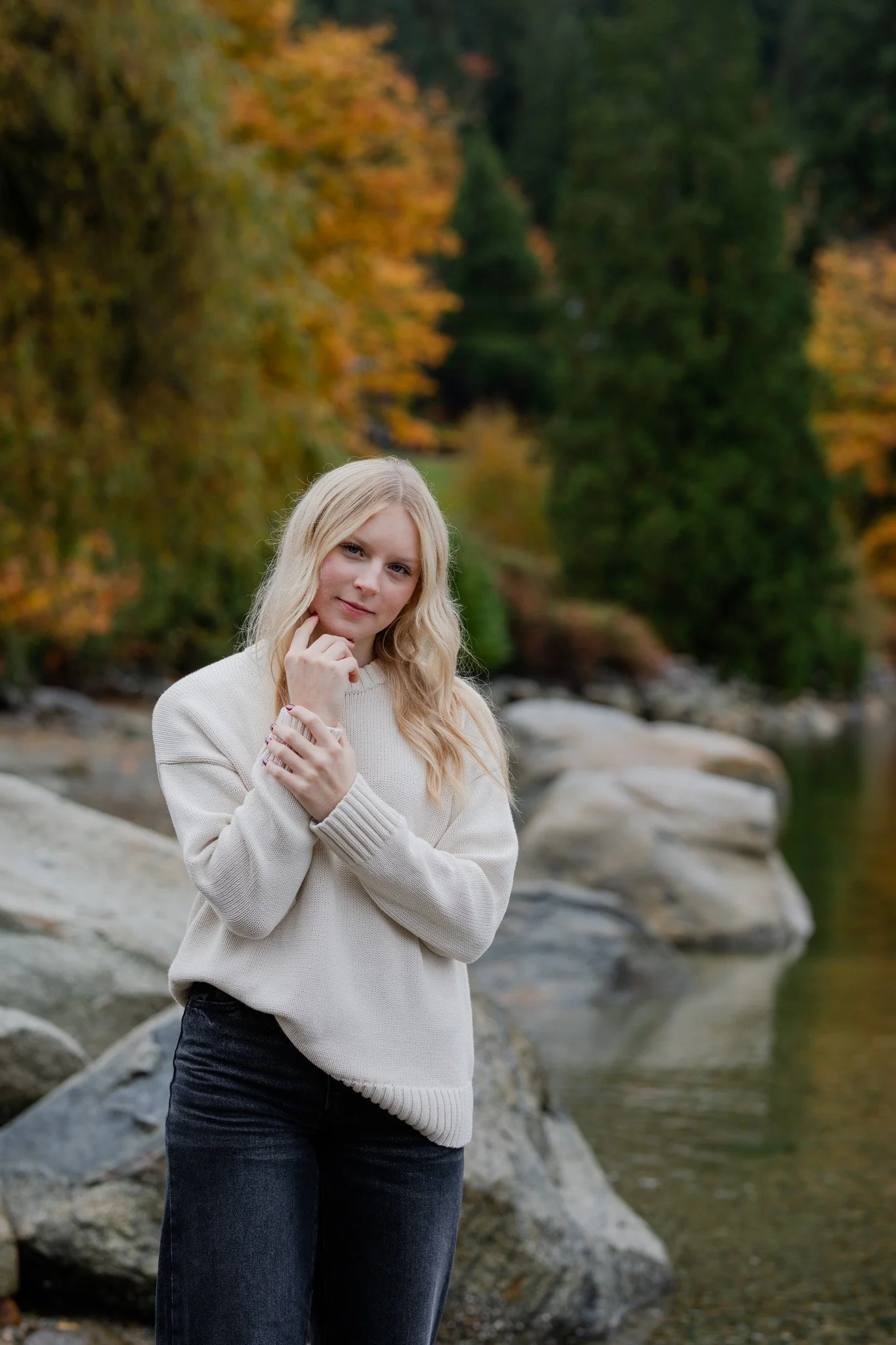 A blonde woman in a cream sweater and black jeans stands near a river with rocks, surrounded by trees with fall foliage.