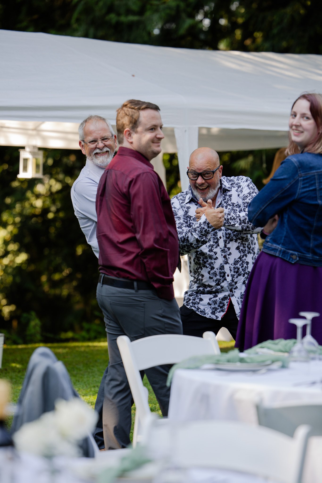 Four adults greeting each other outdoors at a social gathering under a white canopy, with tables and chairs in the foreground and greenery in the background.