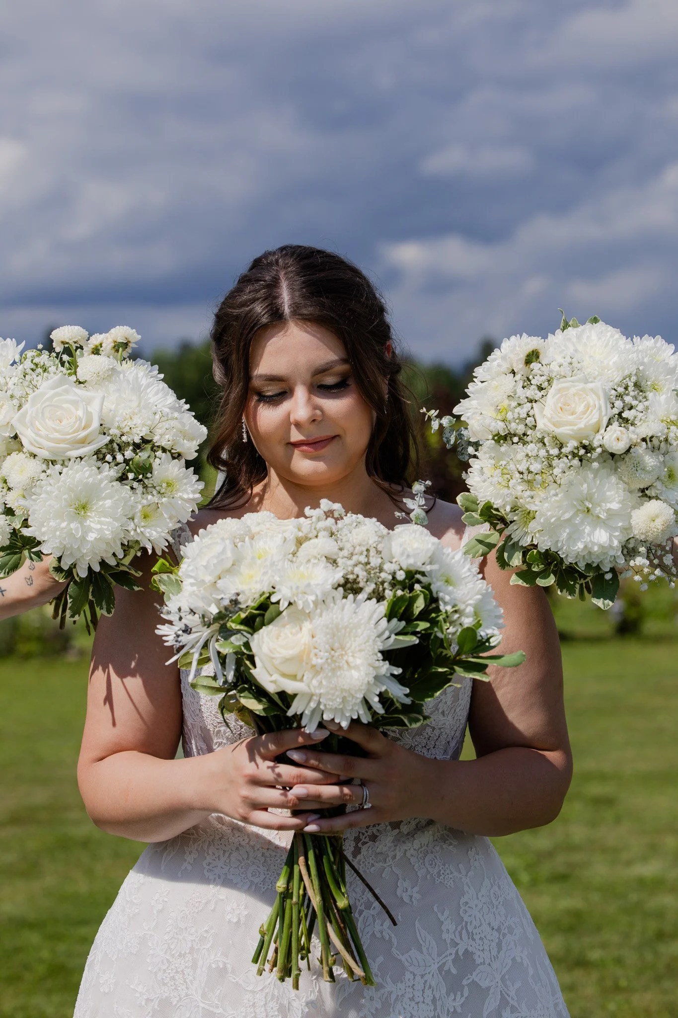 A bride holding a bouquet of white flowers, with more flowers on either side, standing outdoors on a grassy area under a partly cloudy sky.