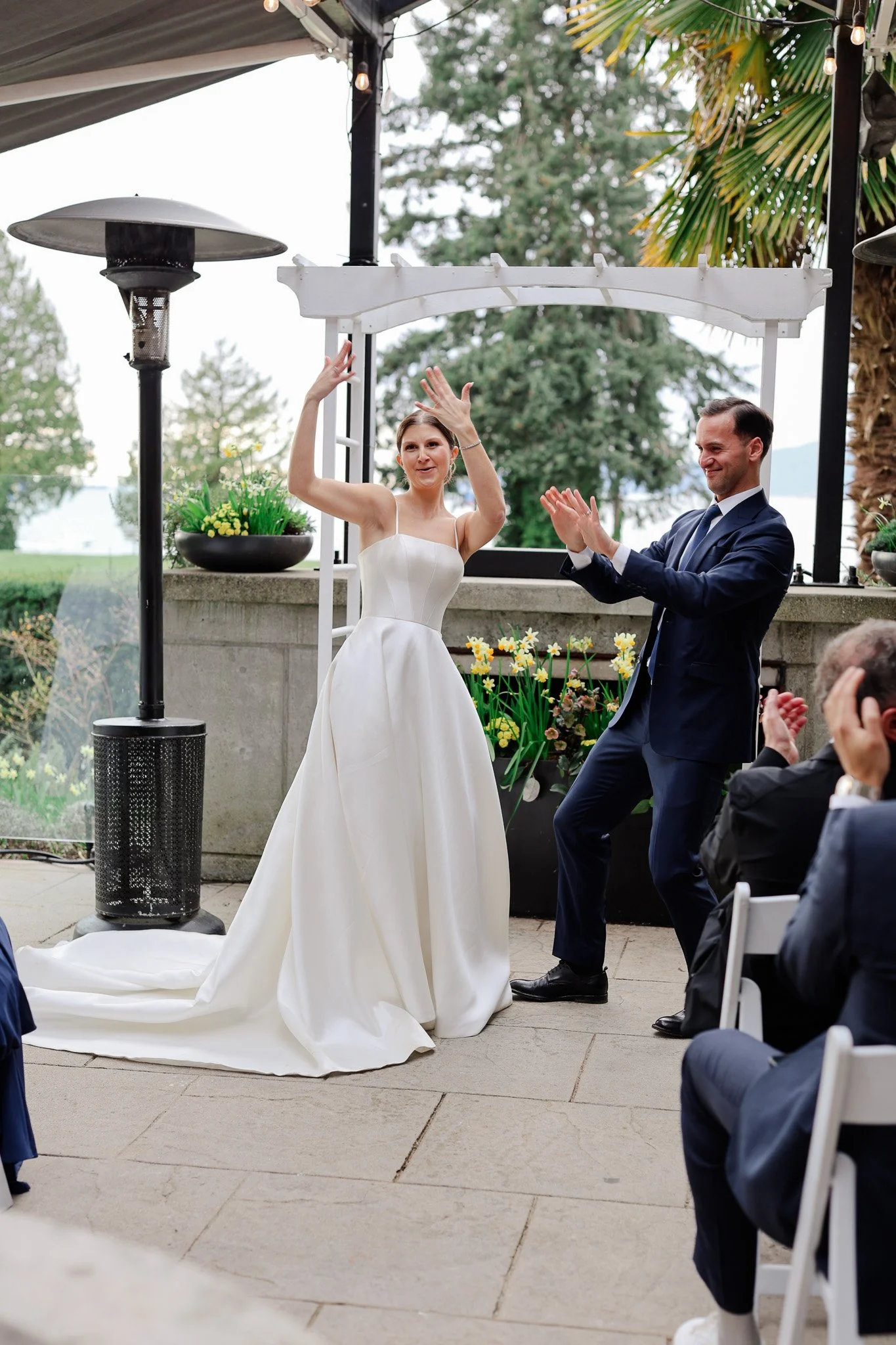 A woman in a white wedding dress and a man in a suit dancing at an outdoor wedding ceremony. They are surrounded by greenery, with chairs and guests visible.