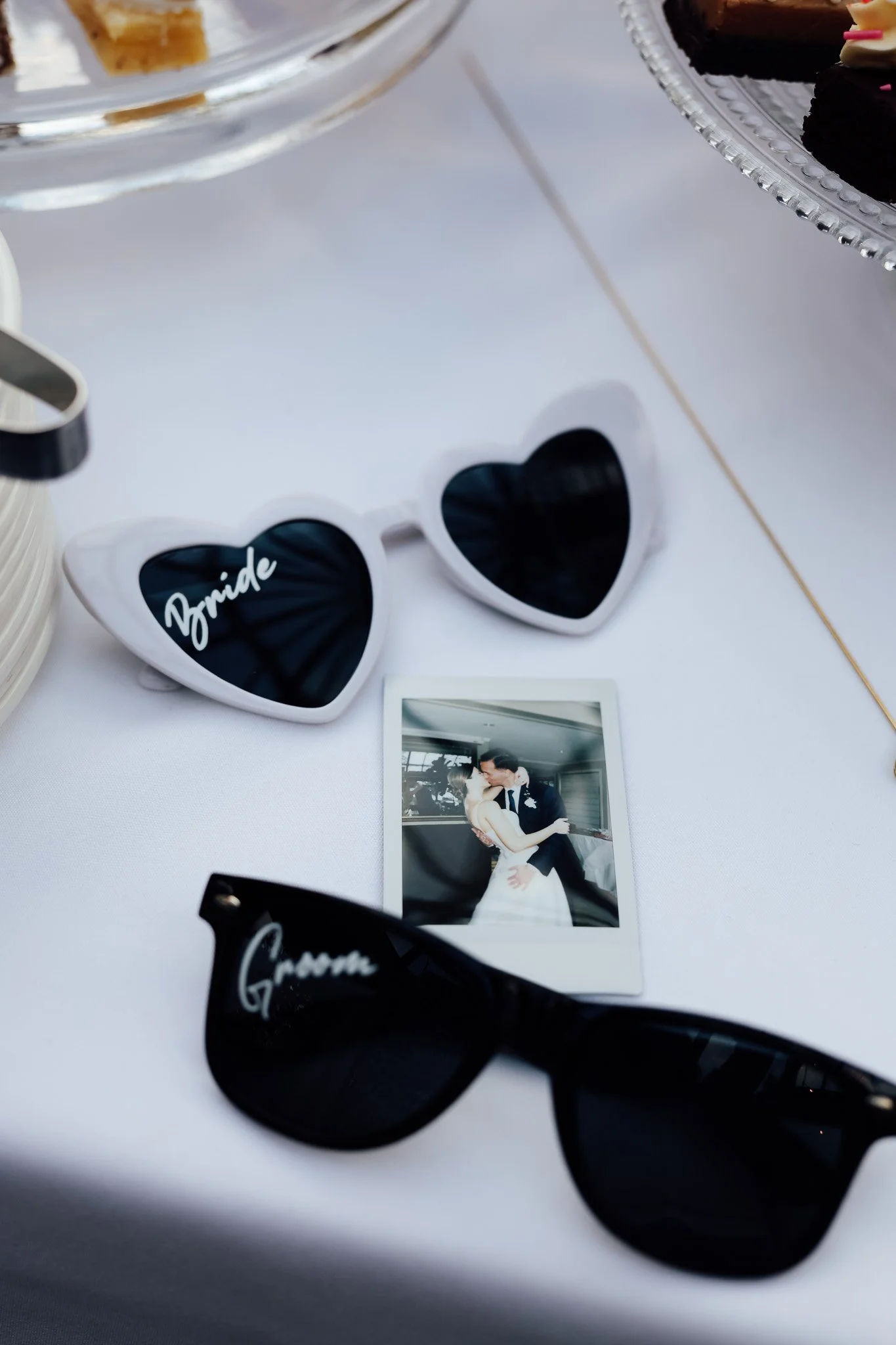 A pair of white heart-shaped sunglasses with the word "bride" written on one lens, a photograph of a couple kissing, and some plates and glasses on a table.