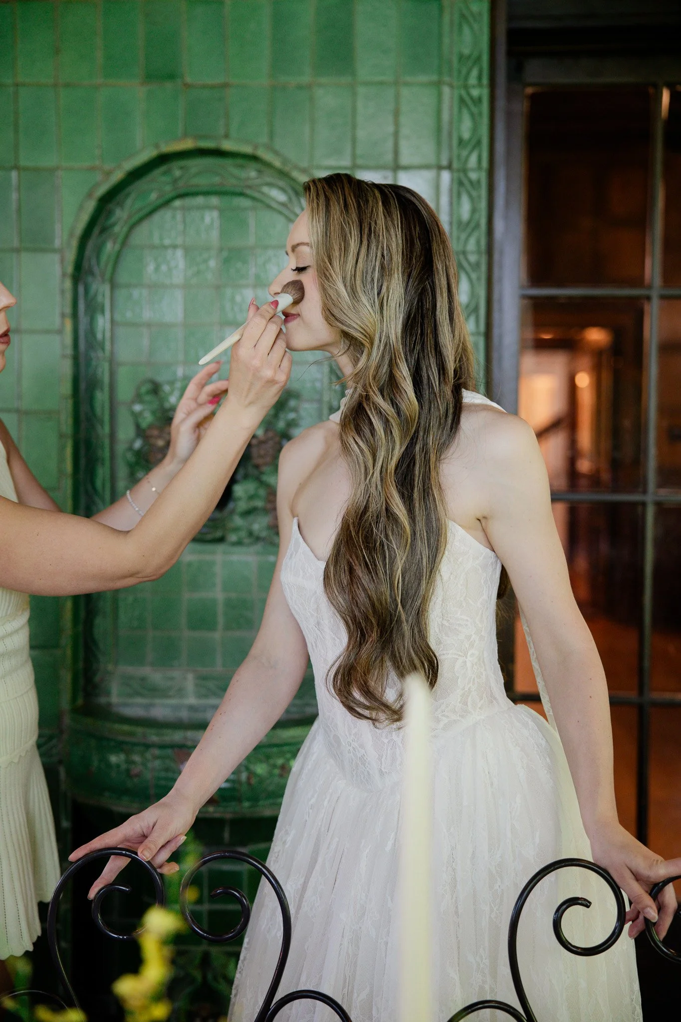 A woman in a white lace dress sits at a table, having makeup applied by another person in a yellow dress. The scene is set in a green-tiled room with a window in the background.