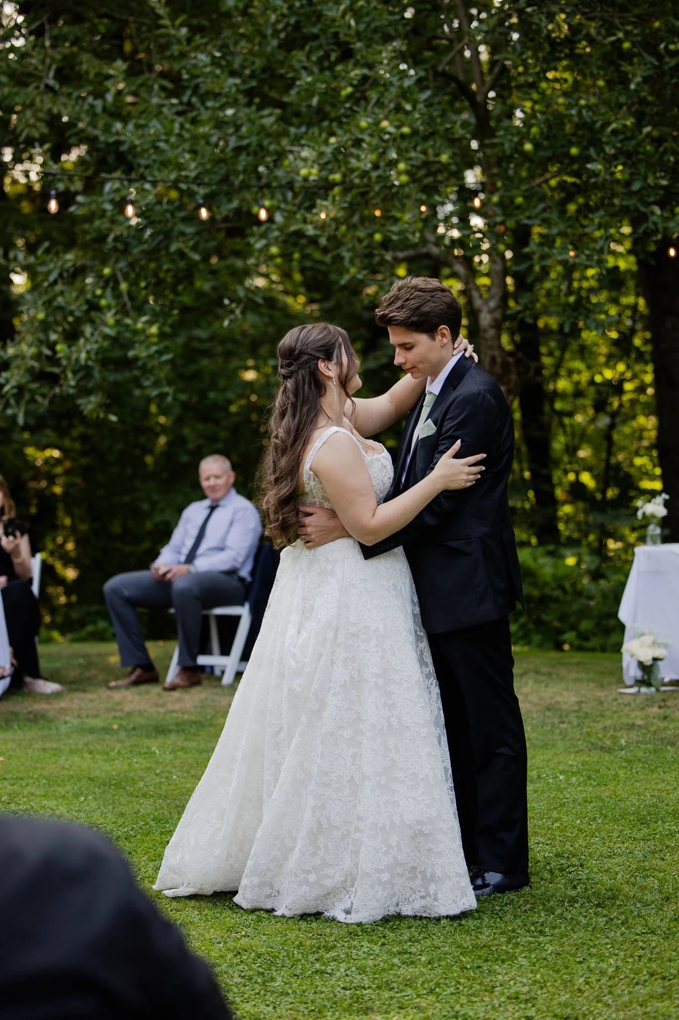 A bride and groom embrace during an outdoor wedding ceremony in a garden, with justices in the background.
