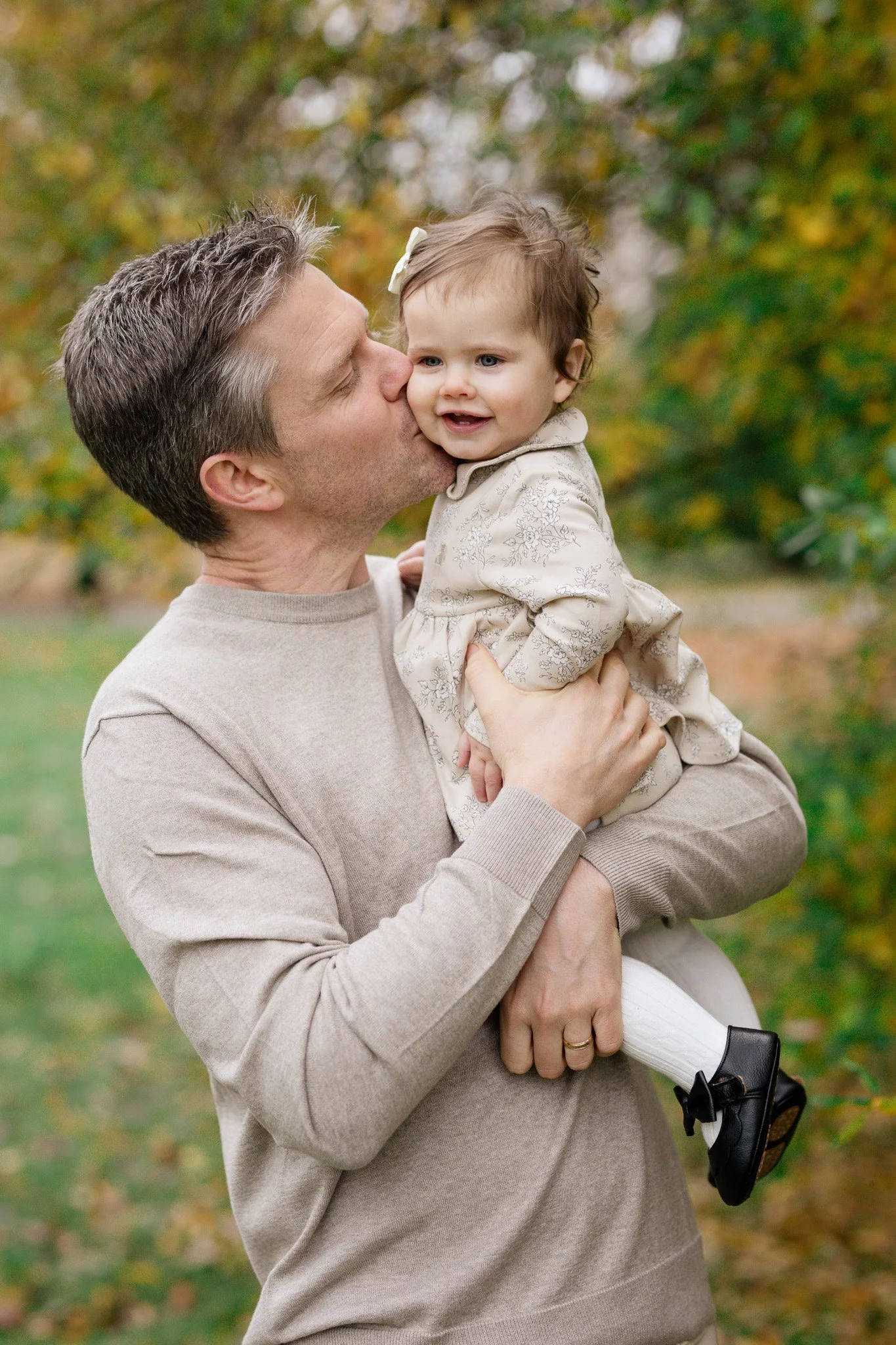 A man holding a young girl outdoors in a park with autumn foliage, the man kisses the girl's cheek.