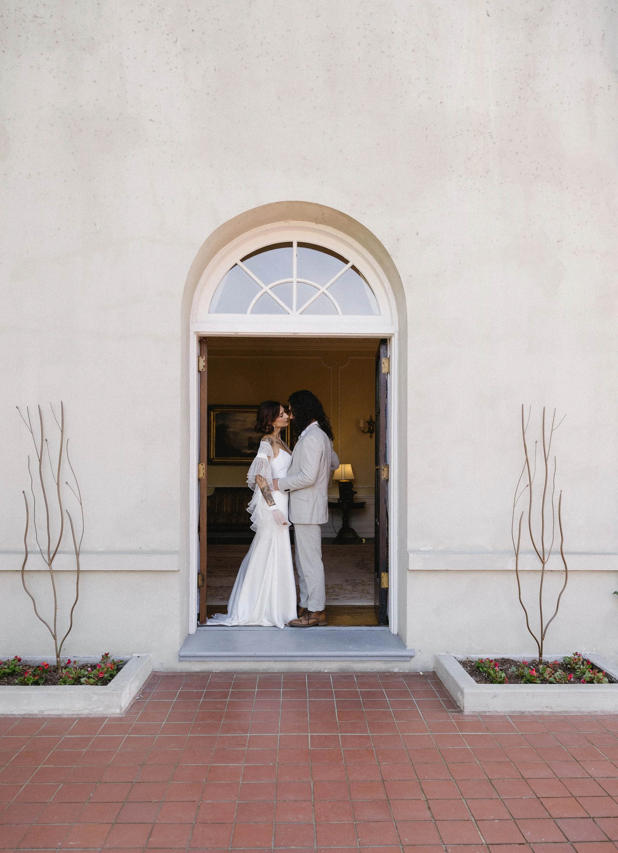 A bride and groom embrace under cherry blossom trees in a park. The bride wears a white wedding gown holding a bouquet, and the groom wears a dark suit.