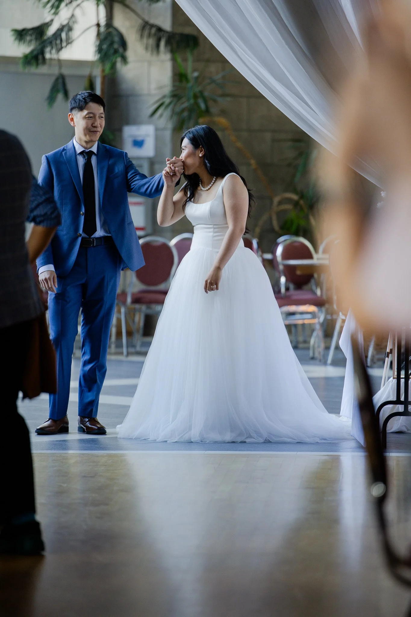 A woman in a white wedding dress kisses a man's hand while he dressed in a blue suit at a wedding reception.