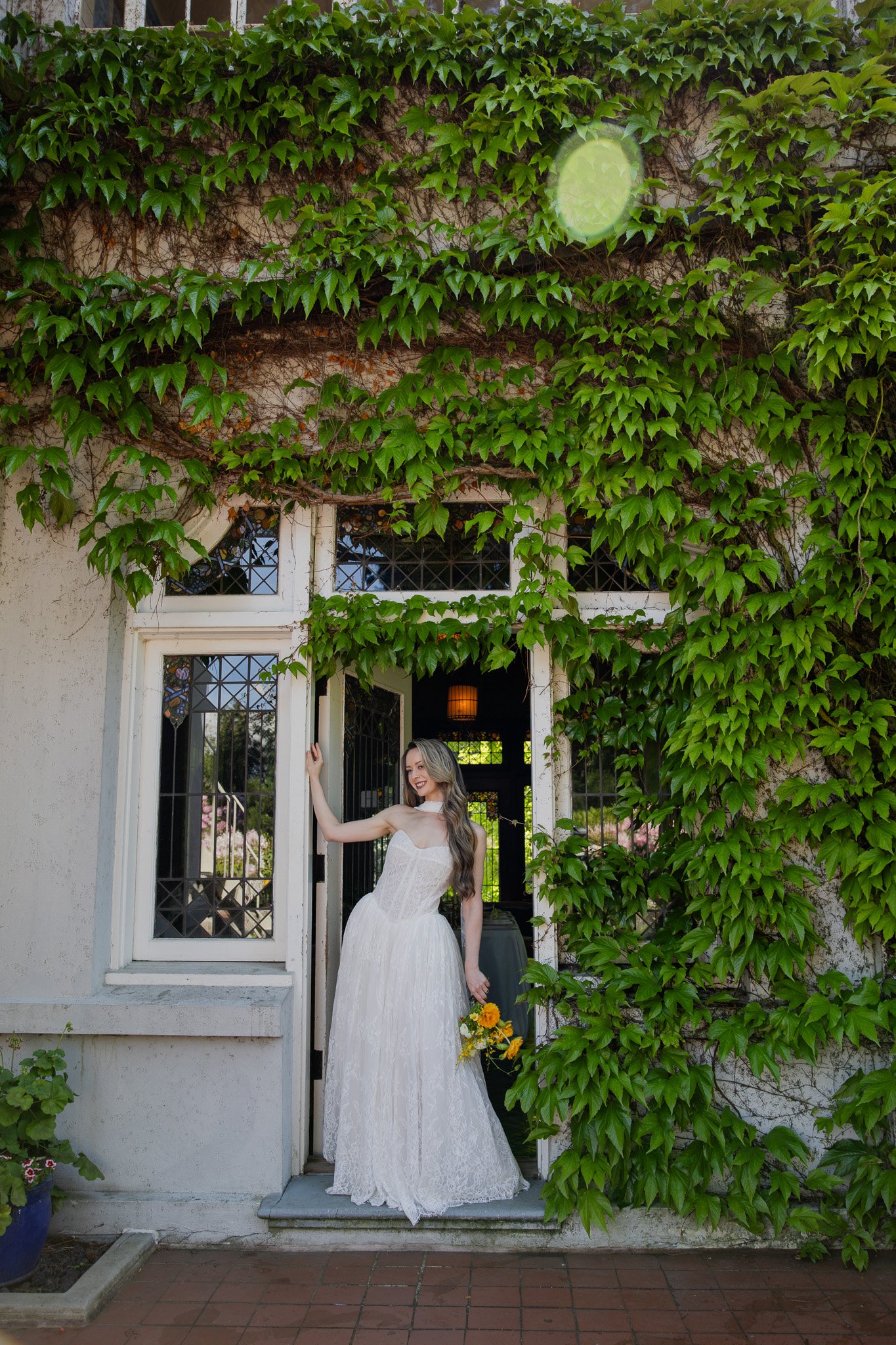 A woman in a white wedding dress standing on a doorstep, holding a bouquet of yellow flowers, with a smiling expression. Green ivy covers the wall around her, with open door and window frames and some pink flowers in the background.