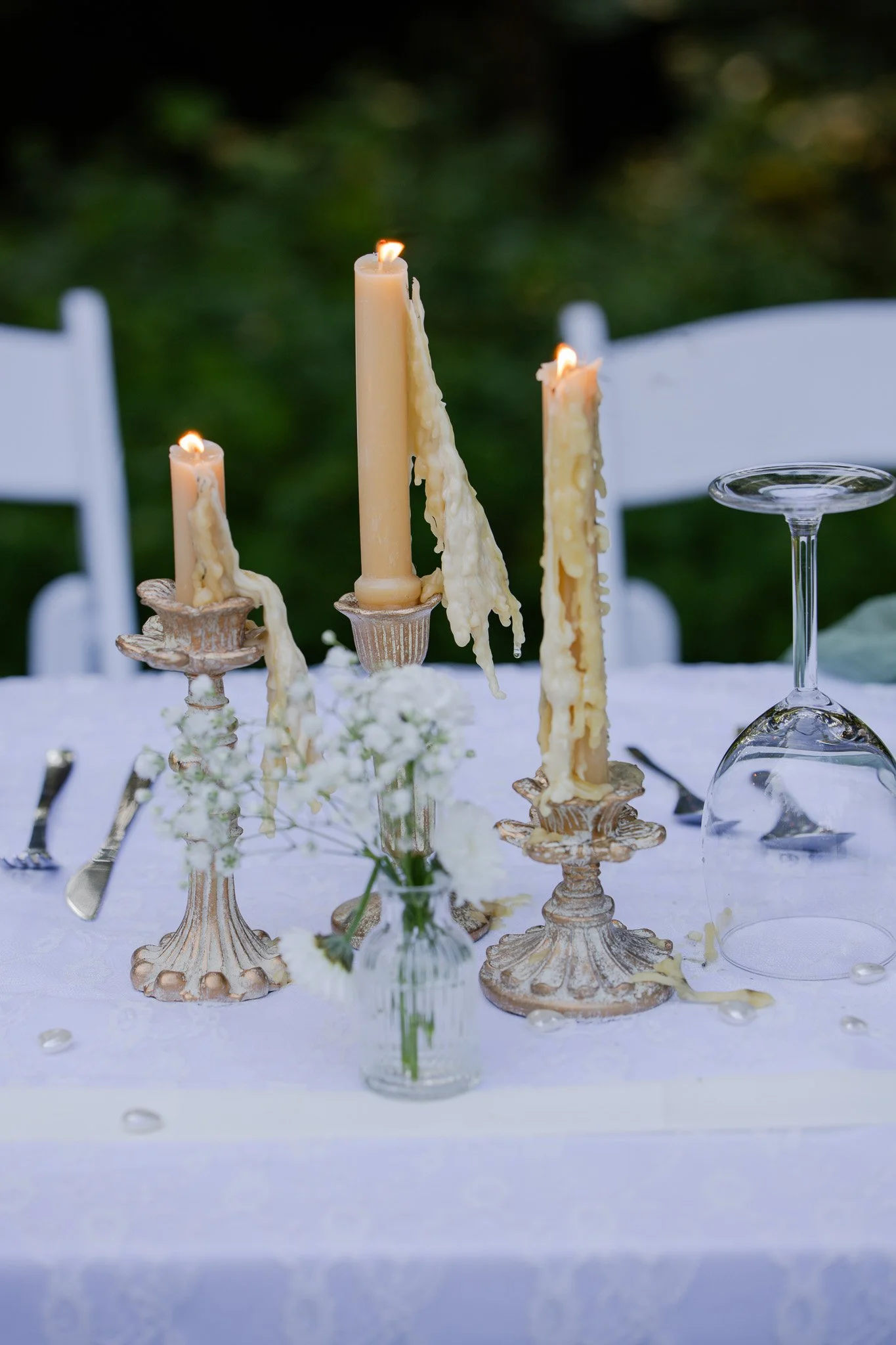 A table decorated with beige candles in ornate candle holders, some of which are melting and dripping wax, with a floral centerpiece and empty fine glassware, set outdoors with a green blurred background.