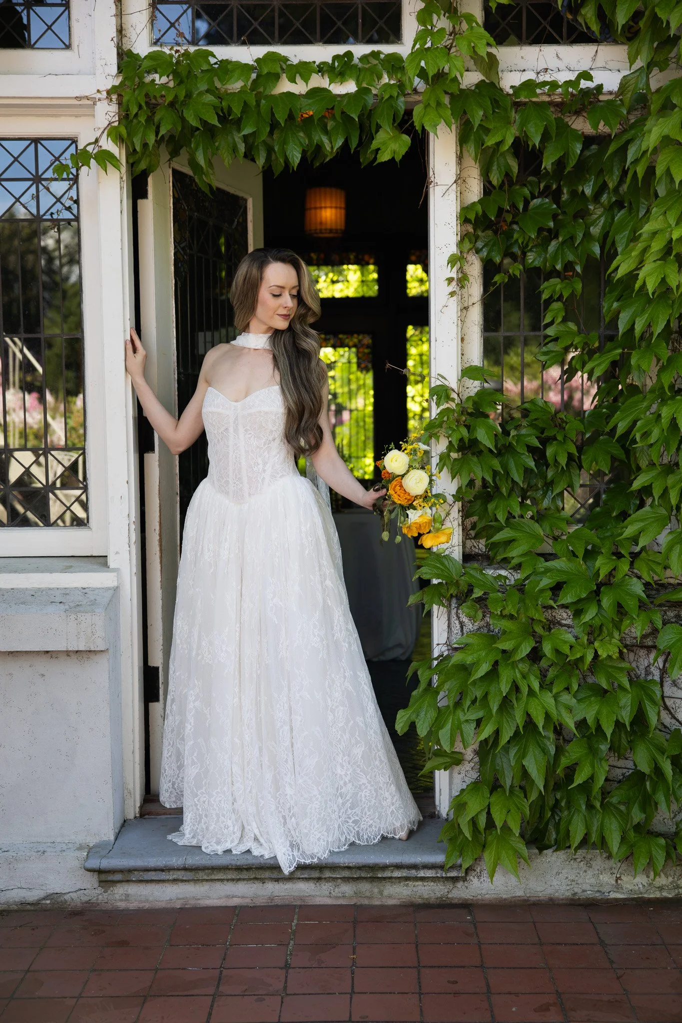 A young woman in a white wedding dress holding a bouquet of yellow and orange flowers, standing at the entrance of a house decorated with green ivy.