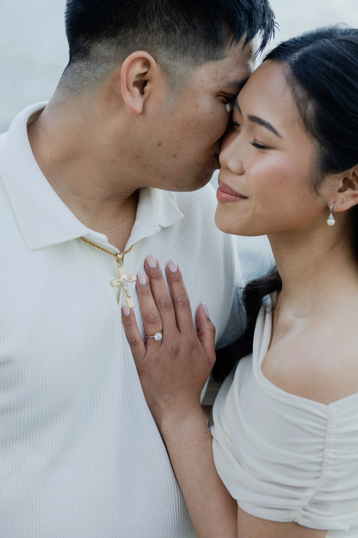 A man and woman are close together, touching foreheads and smiling softly. The man is wearing a white shirt and gold cross necklace, and the woman is wearing a white top with puffed sleeves and pearl earrings.