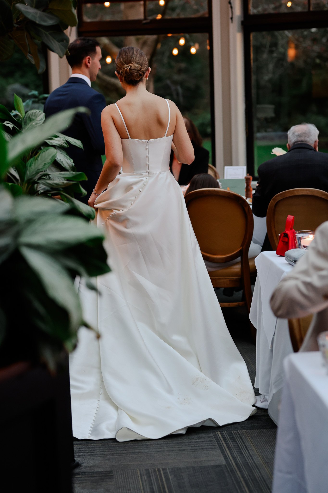 A bride in a white wedding gown with a full skirt and spaghetti straps stands in an indoor venue, facing away. A groom in a dark suit stands nearby, with guests seated at tables.