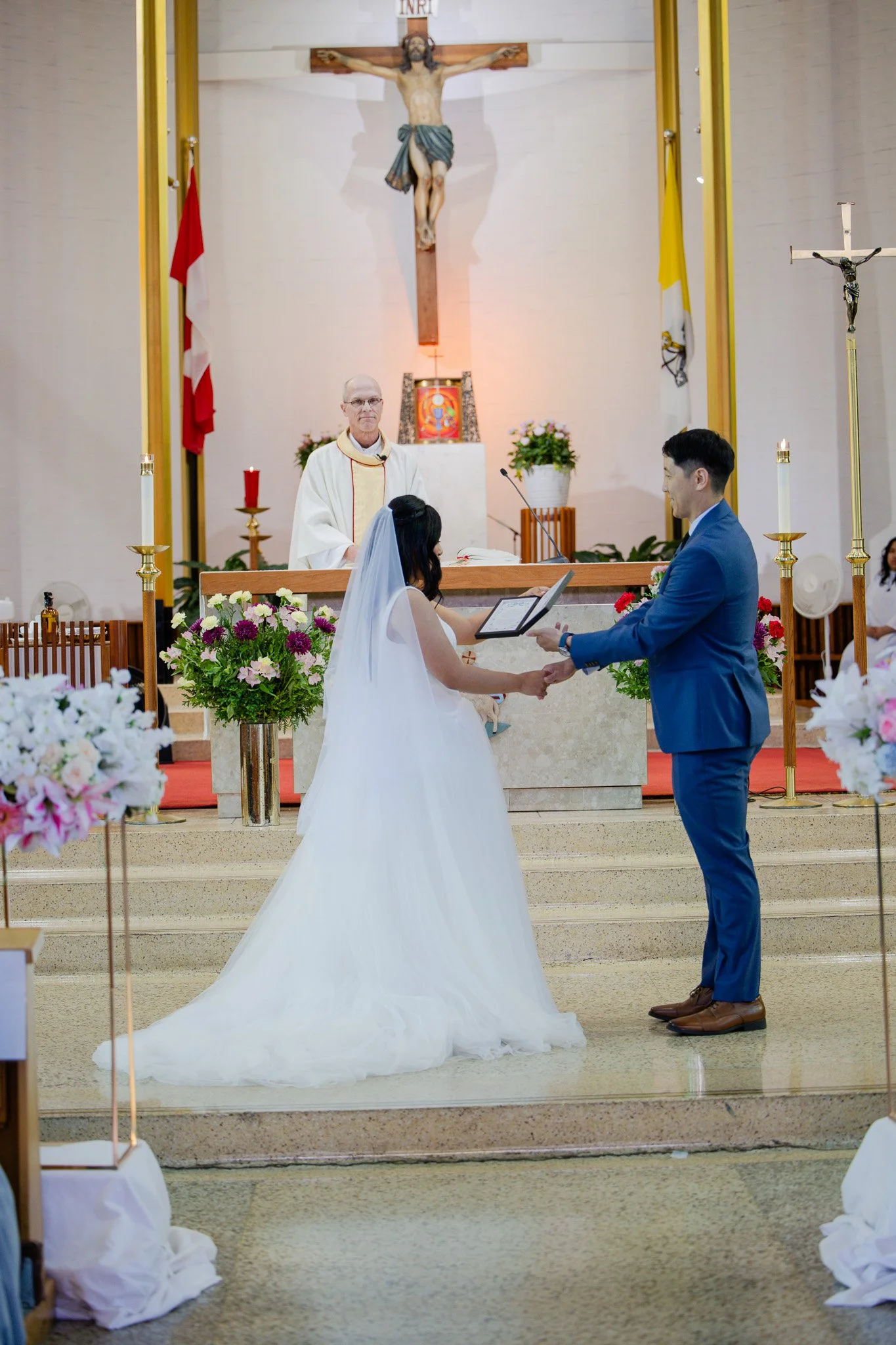 A wedding ceremony taking place in a church with a bride in a white gown and veil, a groom in a blue suit, and a priest or minister standing behind the altar. The bride and groom are holding hands, and the priest is wearing white robes with a gold st