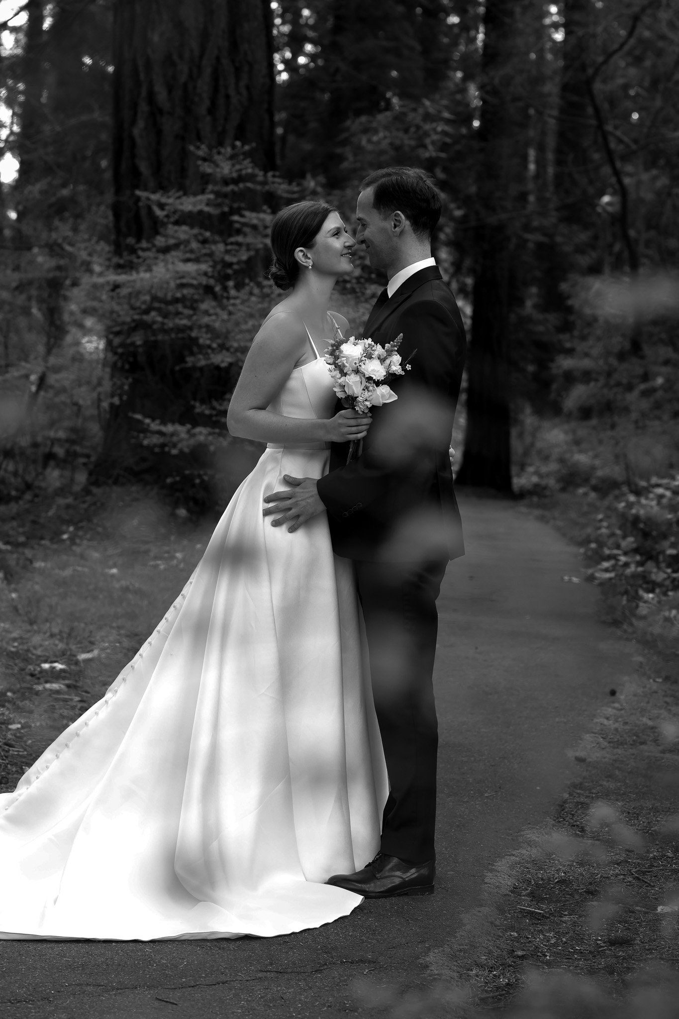 A black-and-white photograph of a bride and groom standing close together outdoors in a wooded area, smiling and facing each other, with the bride holding a bouquet of flowers.