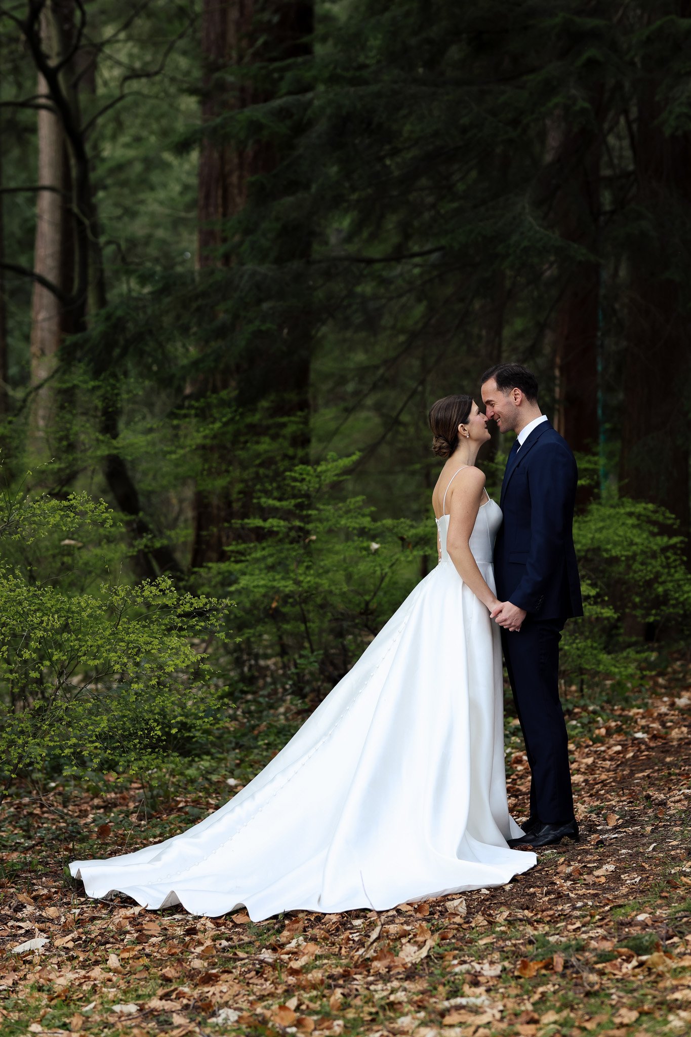 A bride and groom standing close, smiling and touching foreheads in a wooded forest setting.
