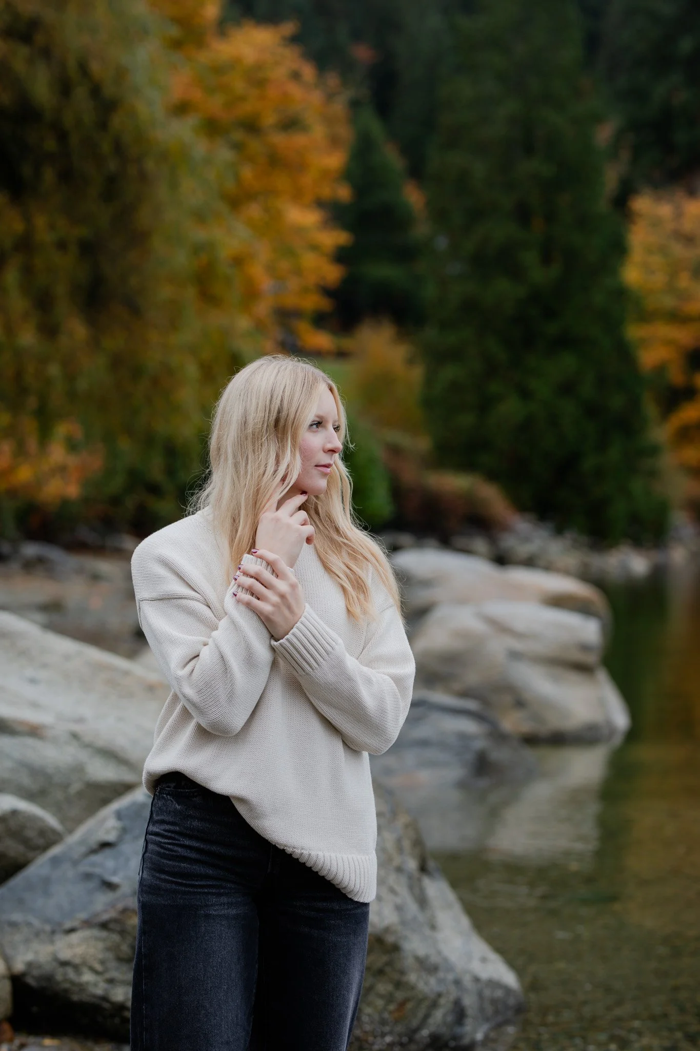 A woman with blonde hair standing outdoors near a river with rocks and trees with autumn foliage in the background.
