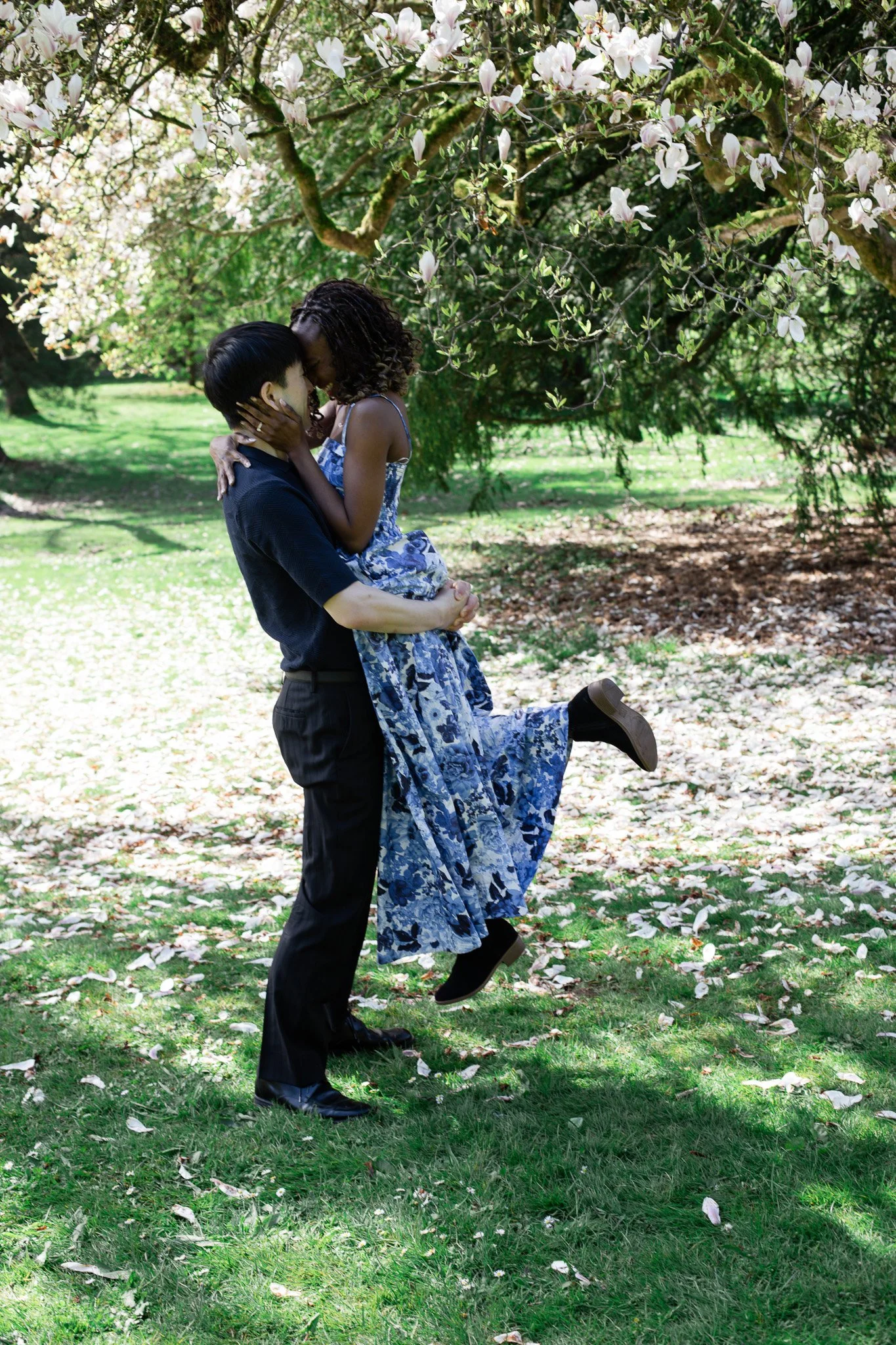 A person in dark clothing is lifting and holding a woman in a blue and white floral dress under a flowering tree, with green grass and fallen petals in the background.