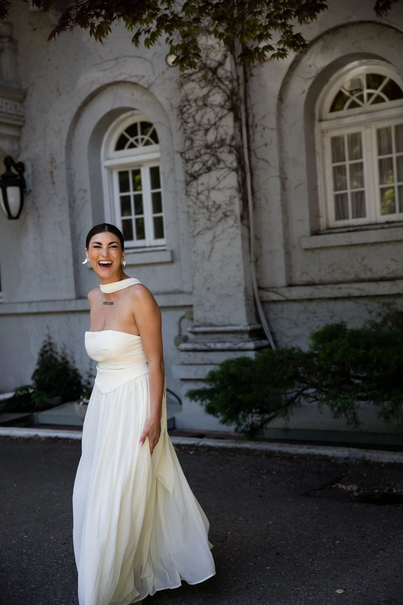 A woman in a white strapless dress with a pleated skirt smiling and standing outside in front of a stone building with arched windows and a vine growing on the wall.