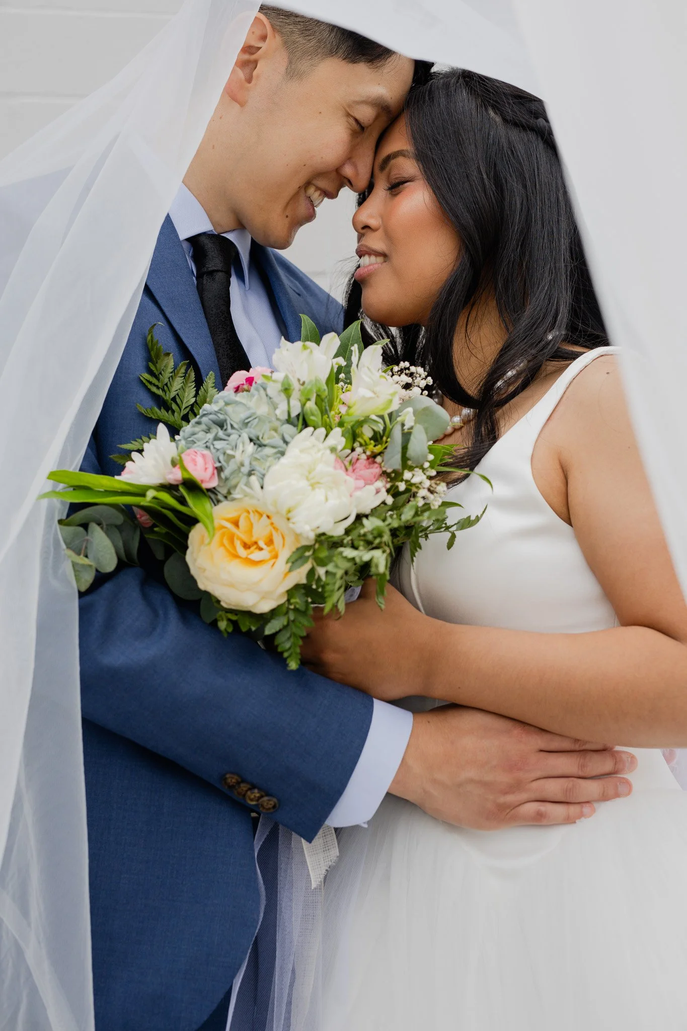 A close-up of a happy couple with foreheads touching, smiling, and holding a bouquet of flowers, under a sheer white fabric, likely at a wedding.