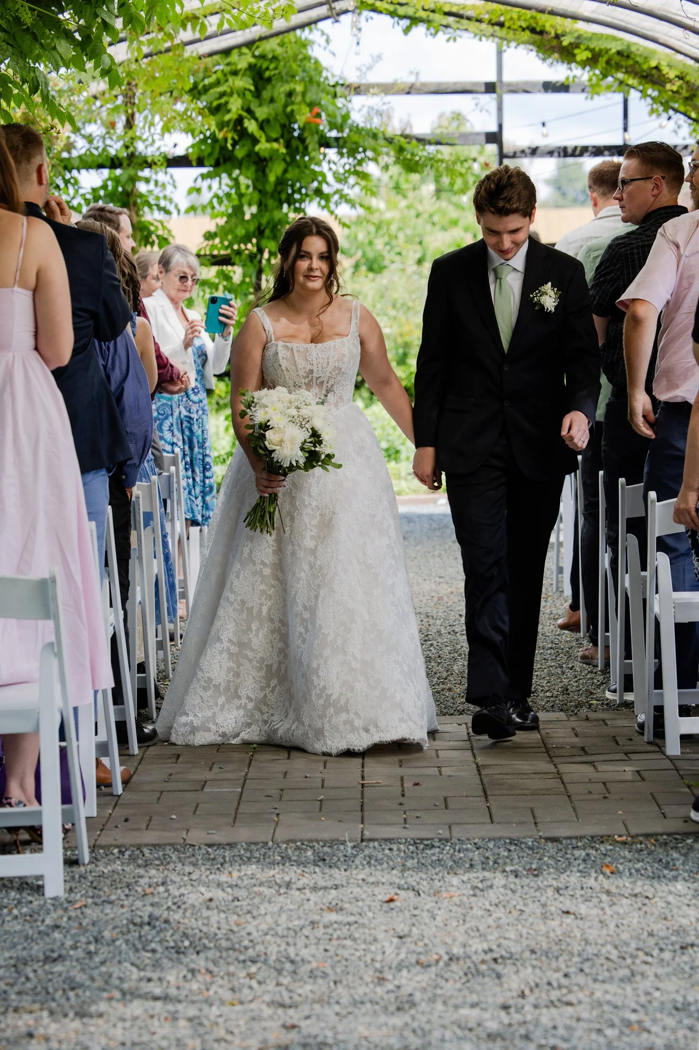 A bride and groom walking down the aisle at an outdoor wedding ceremony, surrounded by seated guests, with greenery and string lights in the background.