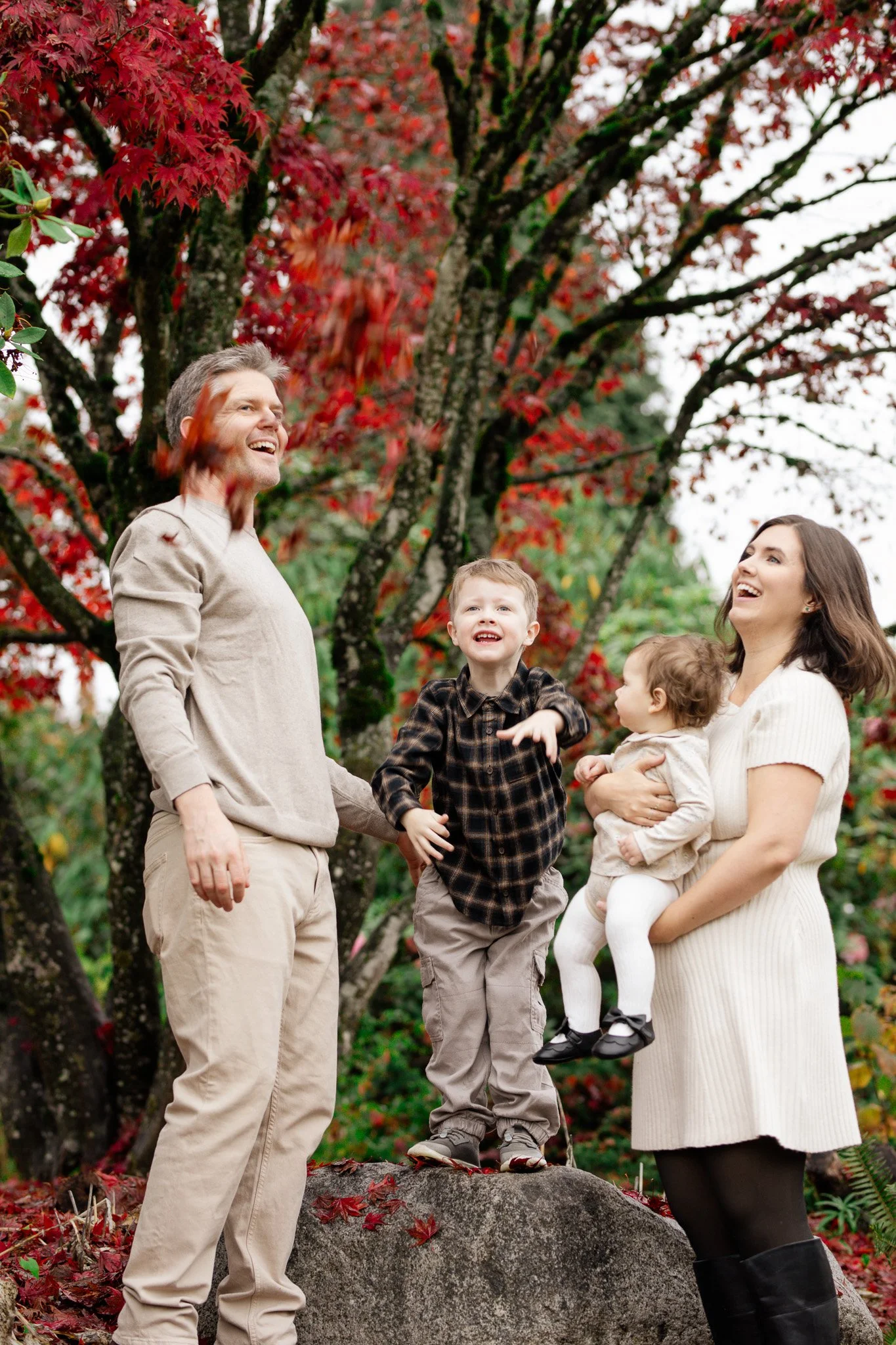 A family of four enjoying a fall day outdoors, with trees with red and orange leaves in the background. The man and woman are smiling, and the young boy is standing on a large rock, smiling and reaching out. The girl is being held by the woman and is