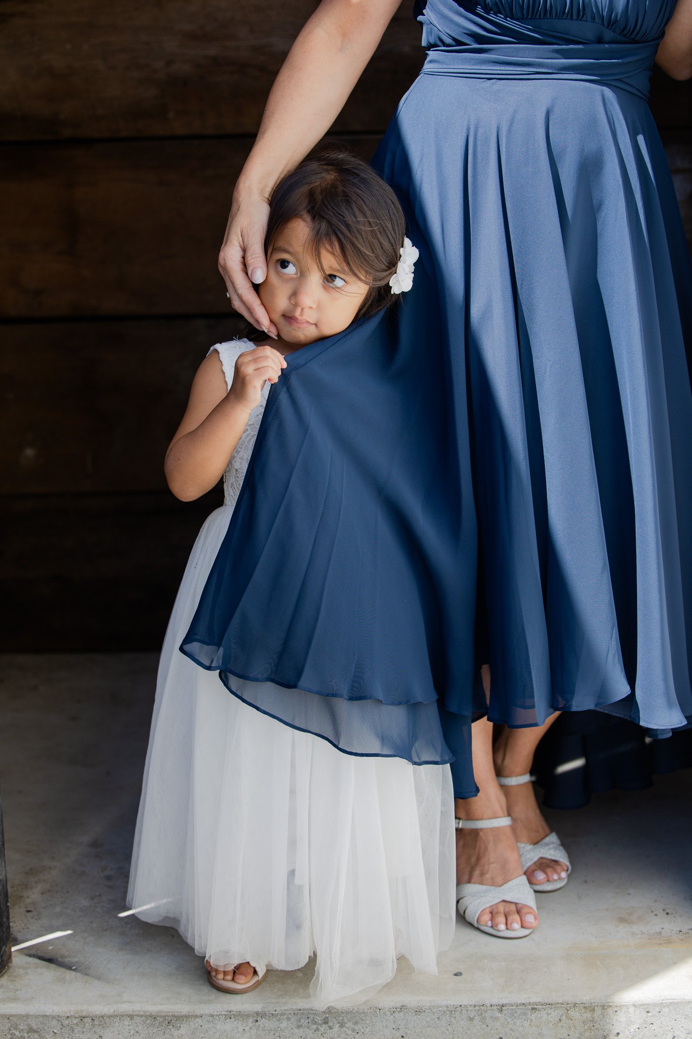 A young girl in a white dress with a floral hair accessory stands next to an adult woman in a blue dress, with the woman placing her hand on the girl's head. The scene appears to be in a formal setting, possibly a wedding or special event, with other