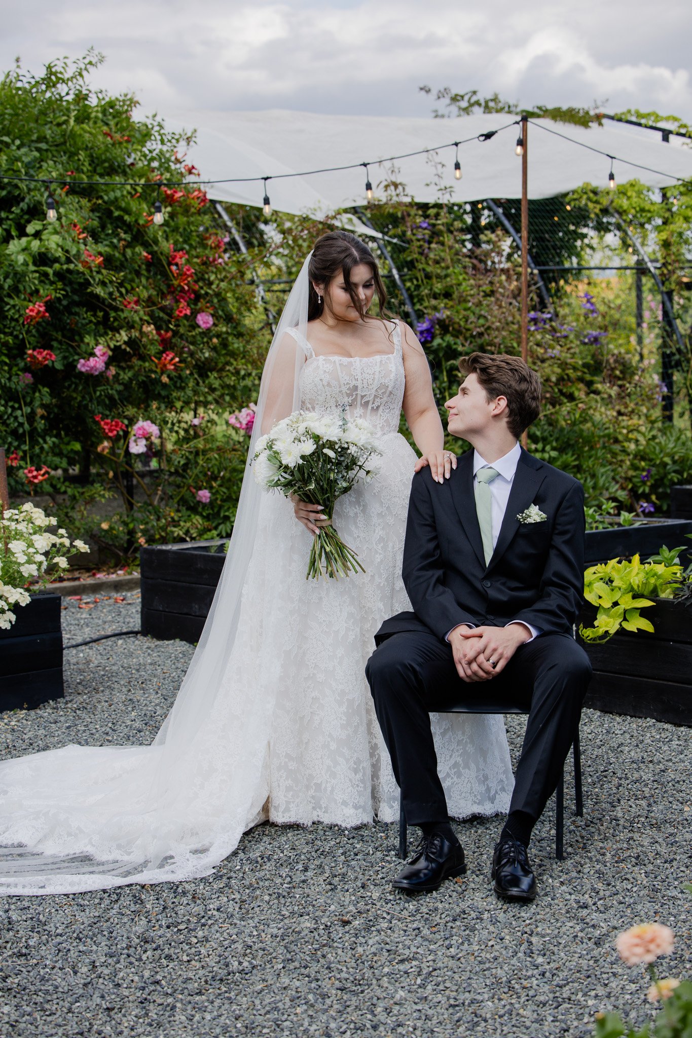 A bride in a white wedding dress and veil holding a bouquet of white flowers stands next to a groom in a black suit, seated outdoors in a garden with colorful flowers and string lights overhead.