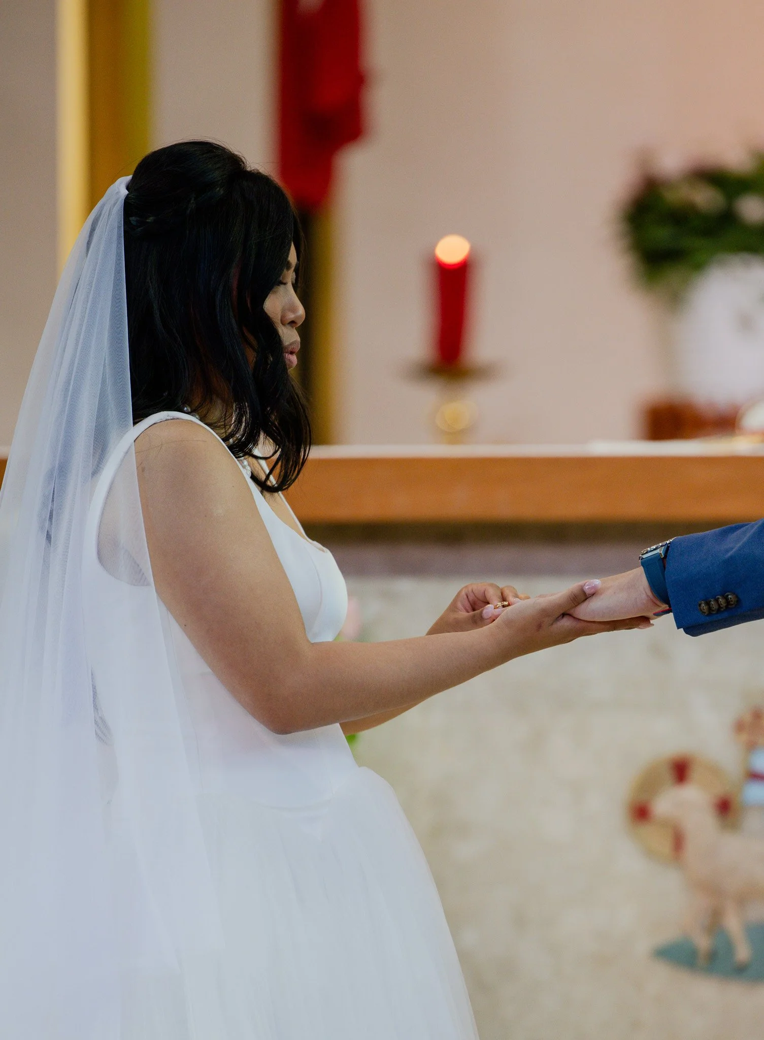 A woman in a white dress with a veil, likely a bride, holding hands with a man dressed in a blue suit during a ceremony, possibly a wedding, inside a church or ceremony hall.
