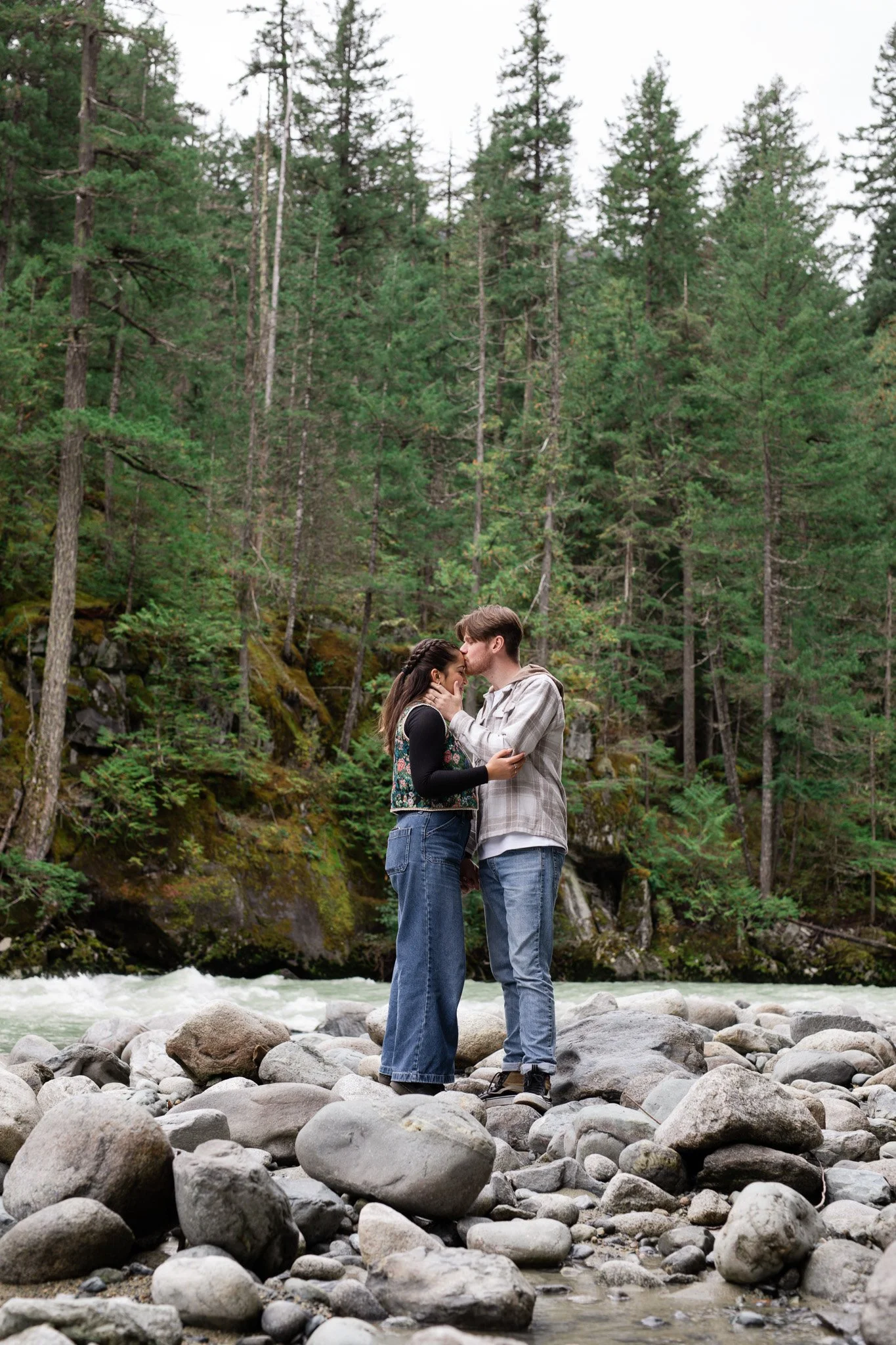 A couple standing on rocks near a river in a forest, sharing a kiss.