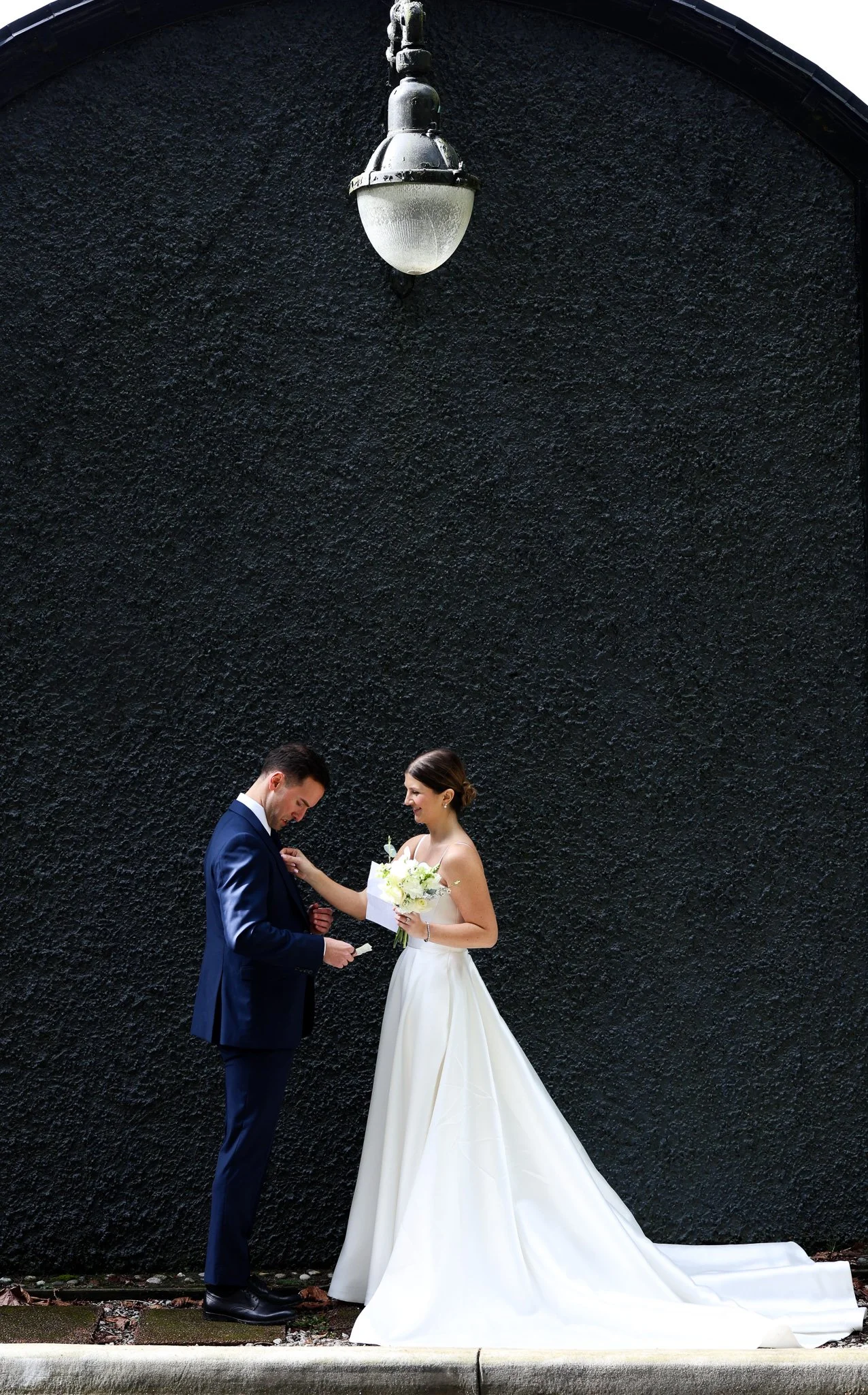 A bride and groom standing on a sidewalk in front of a textured black wall, with the bride holding a bouquet of white flowers and the groom holding a small object, possibly a ring or a piece of paper, during a wedding ceremony or moment.