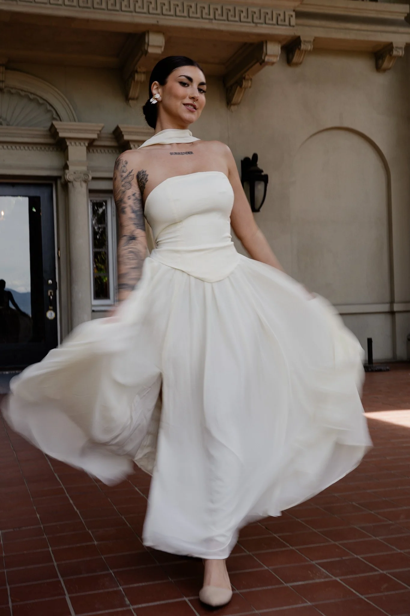 A woman in a strapless white dress twirling, creating a flowing skirt, outside a building with classical architectural details.