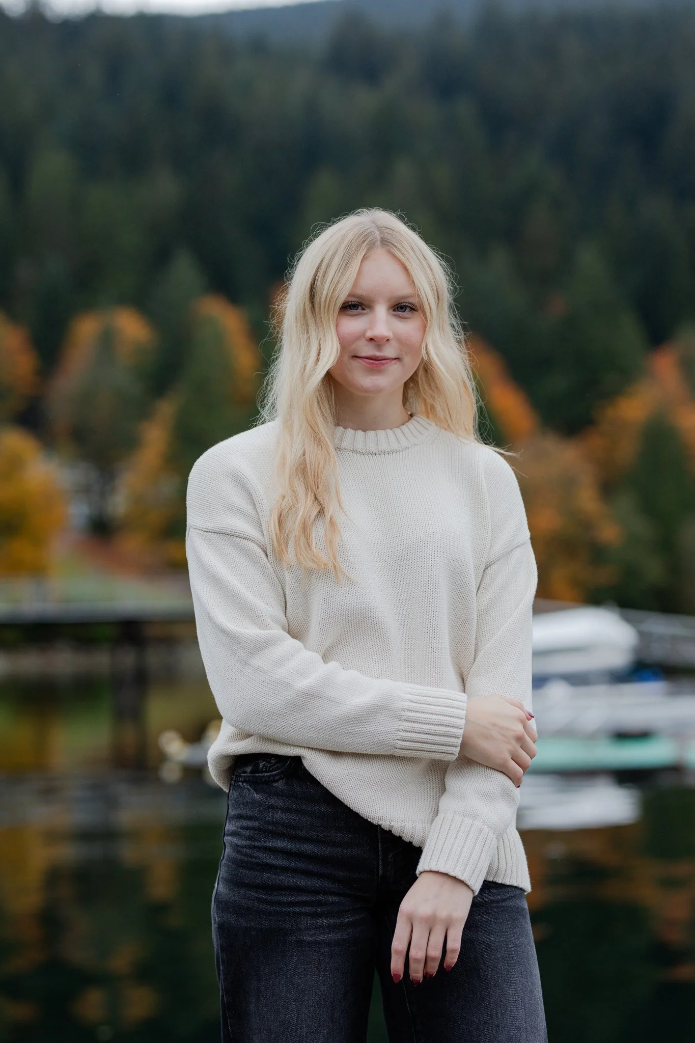 A young woman with blonde wavy hair standing outdoors near a lake, with trees showing autumn colors in the background.