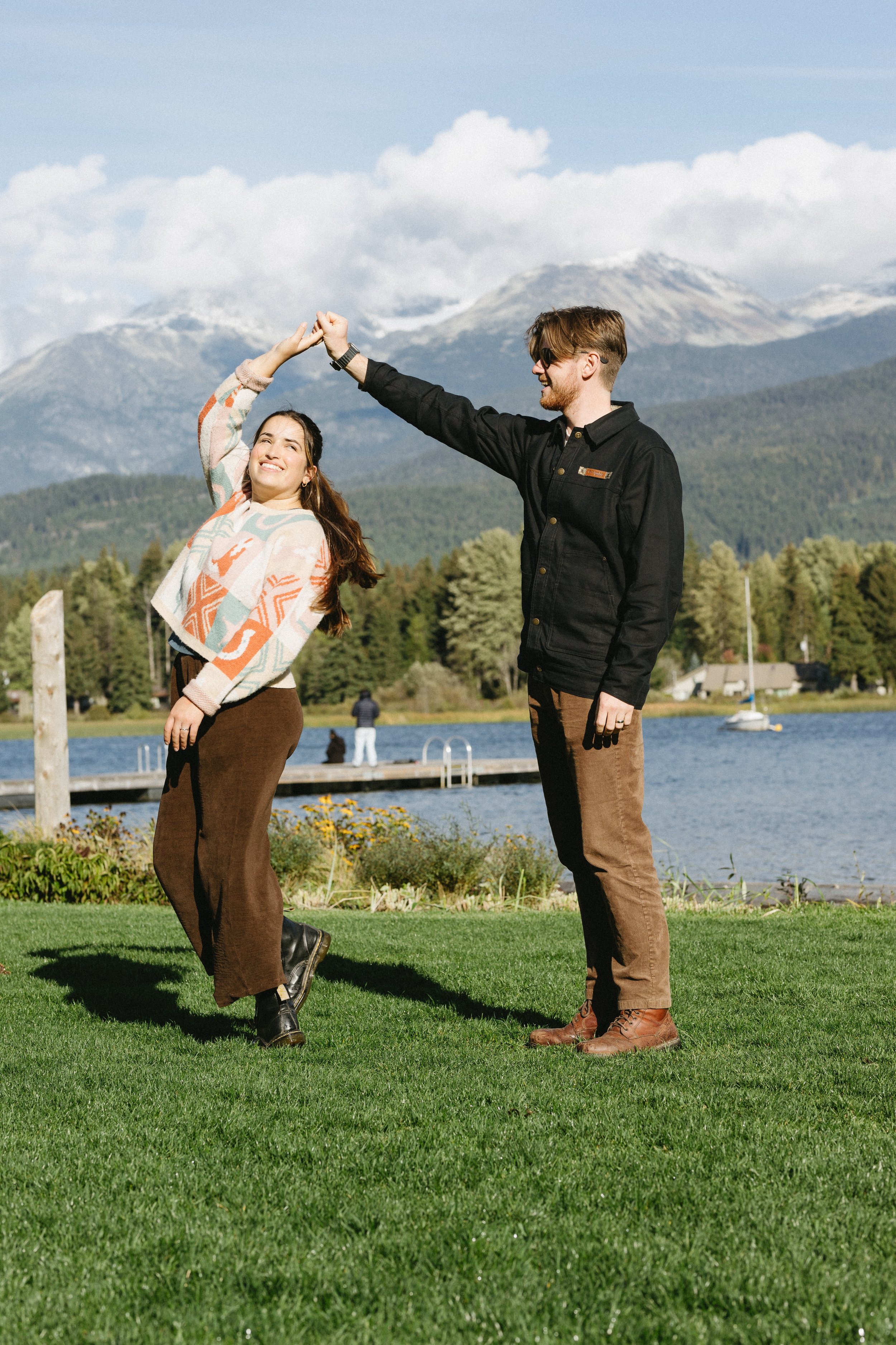 A young couple dancing outdoors on green grass near a lake with mountains in the background on a sunny day.