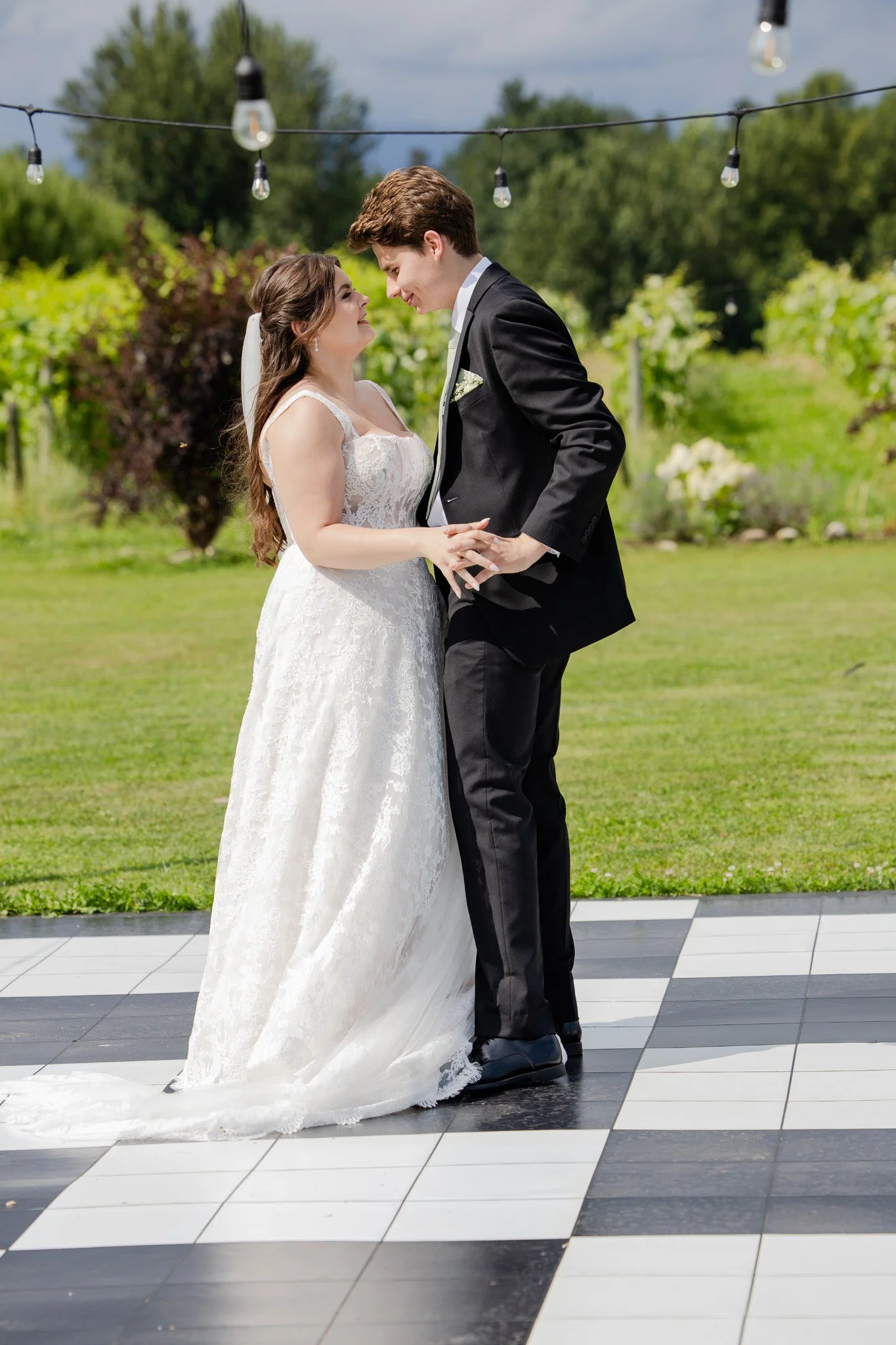 A bride and groom share a dance outdoors on a checkered dance floor during daytime, with string lights hanging above and greenery in the background.
