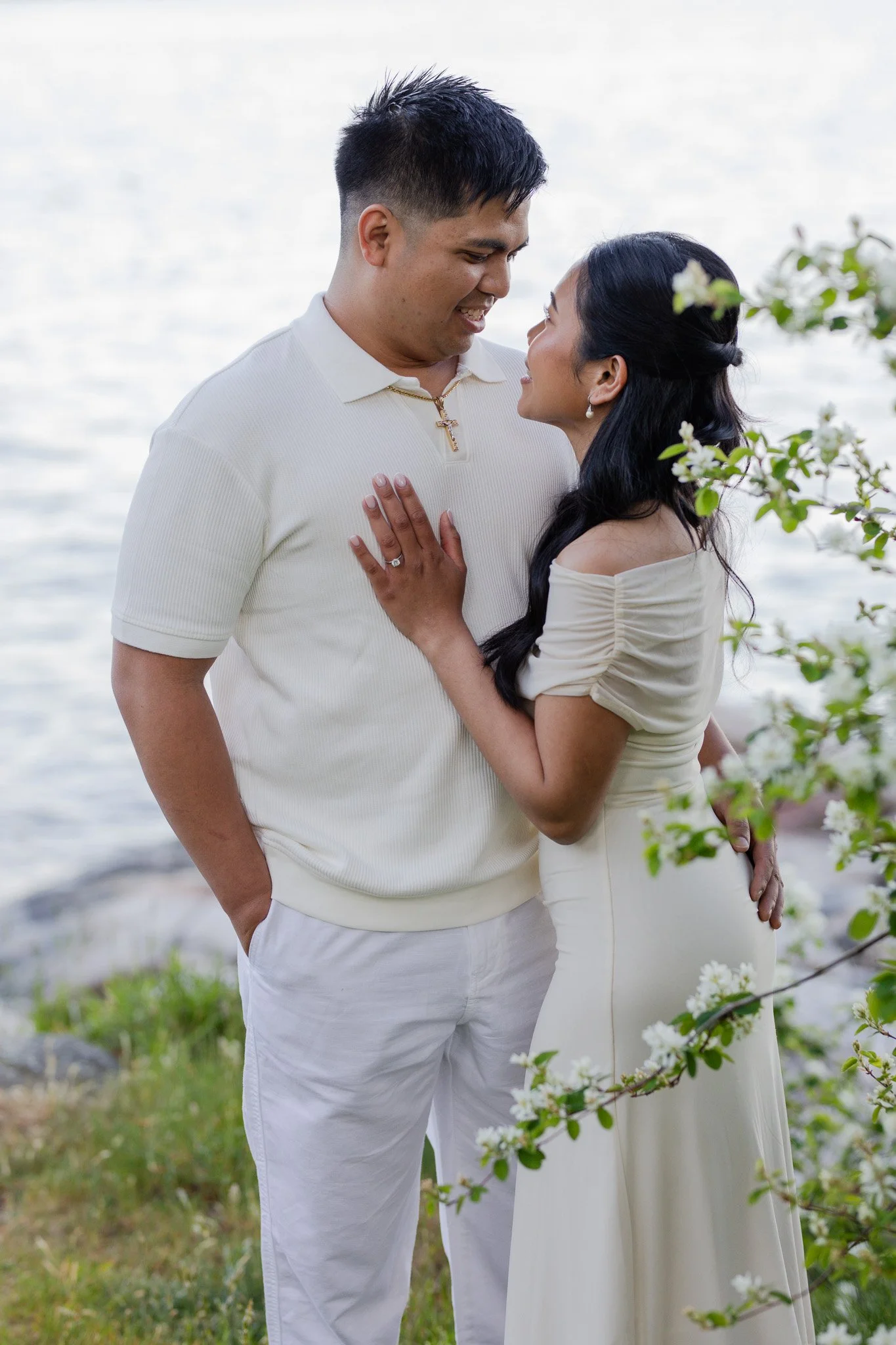 A couple stands close near water, smiling at each other, with the woman holding the man's chest and the man having his hand in his pocket. The woman wears an off-the-shoulder cream dress, and the man is in a white polo and pants. There are white blos