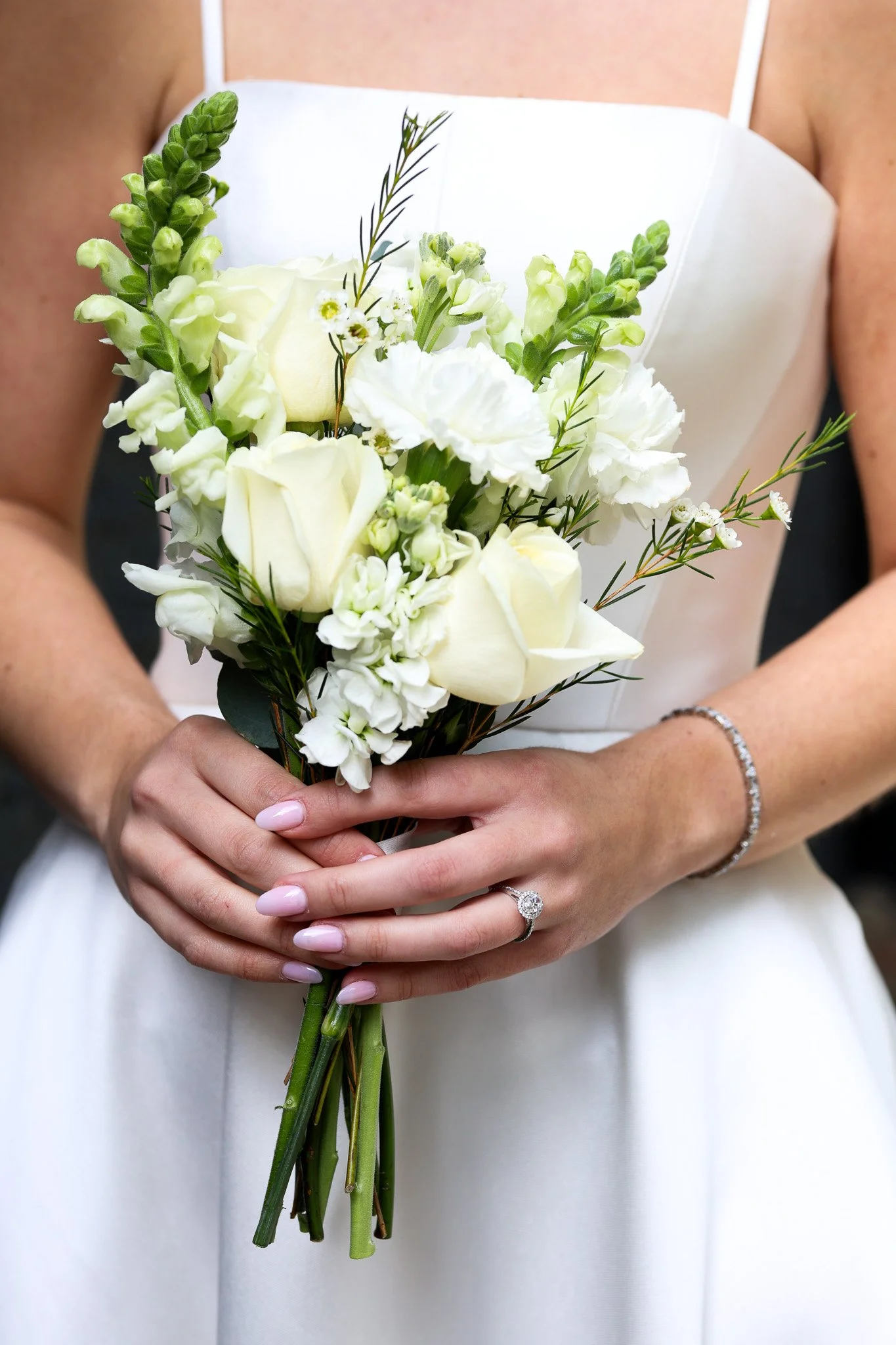 A bride holding a bouquet of white flowers, including roses and lisianthus, with both hands. She is wearing a white dress, an engagement ring, and a bracelet, with her nails painted in a light pink color.
