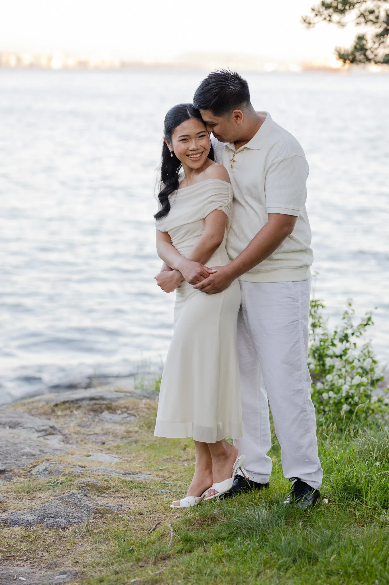 A couple stands close together by a body of water, with the woman smiling and the man whispering in her ear, both dressed in white; the woman wears a off-shoulder dress and white high heels, and the man is in a polo shirt and pants, on a grassy area 