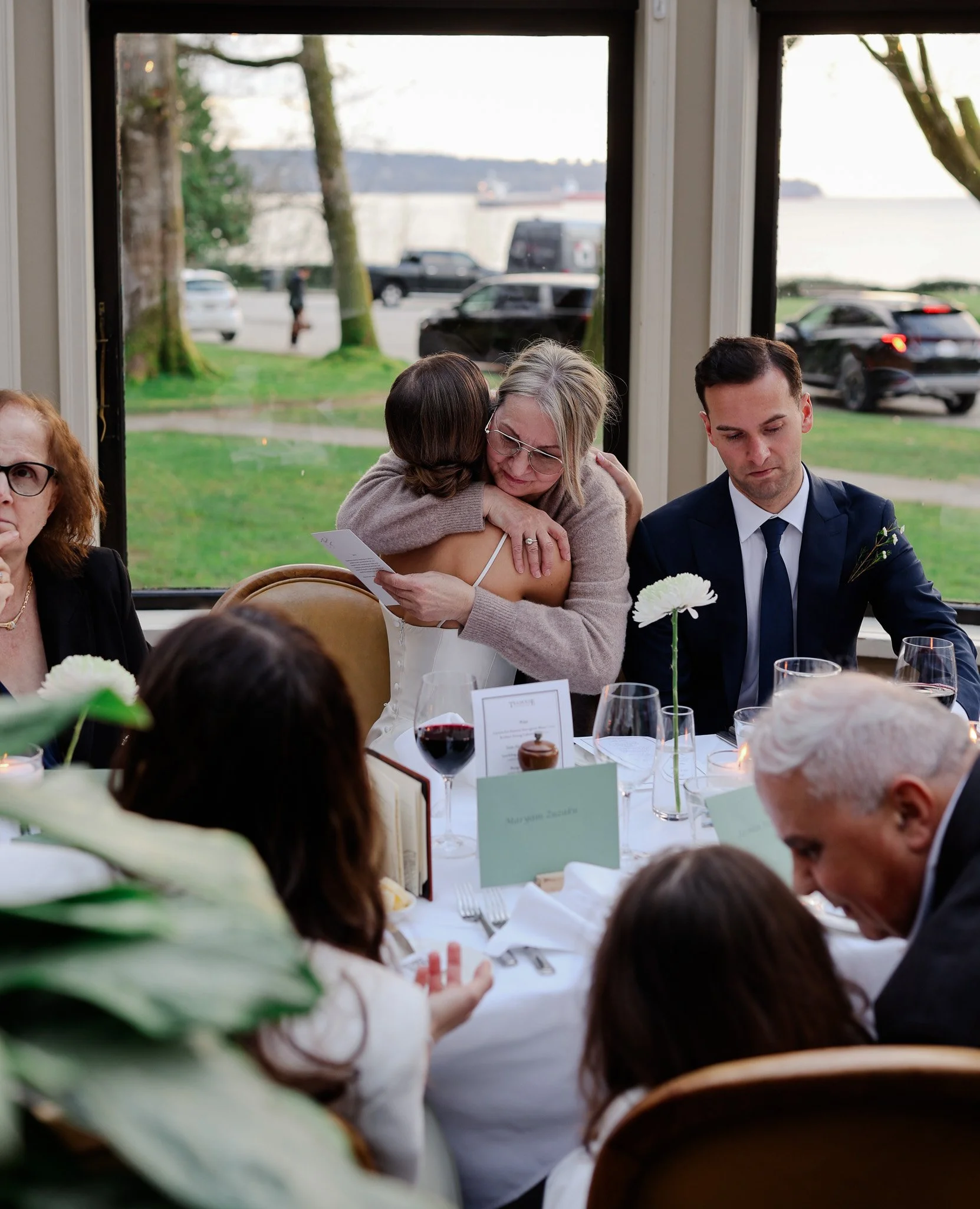 An indoor gathering at a restaurant or banquet hall with large windows showing a parking lot and water outside. A woman with glasses and blonde hair, wearing a beige sweater, is hugging a woman with short brown hair, seen from behind. To the right, a
