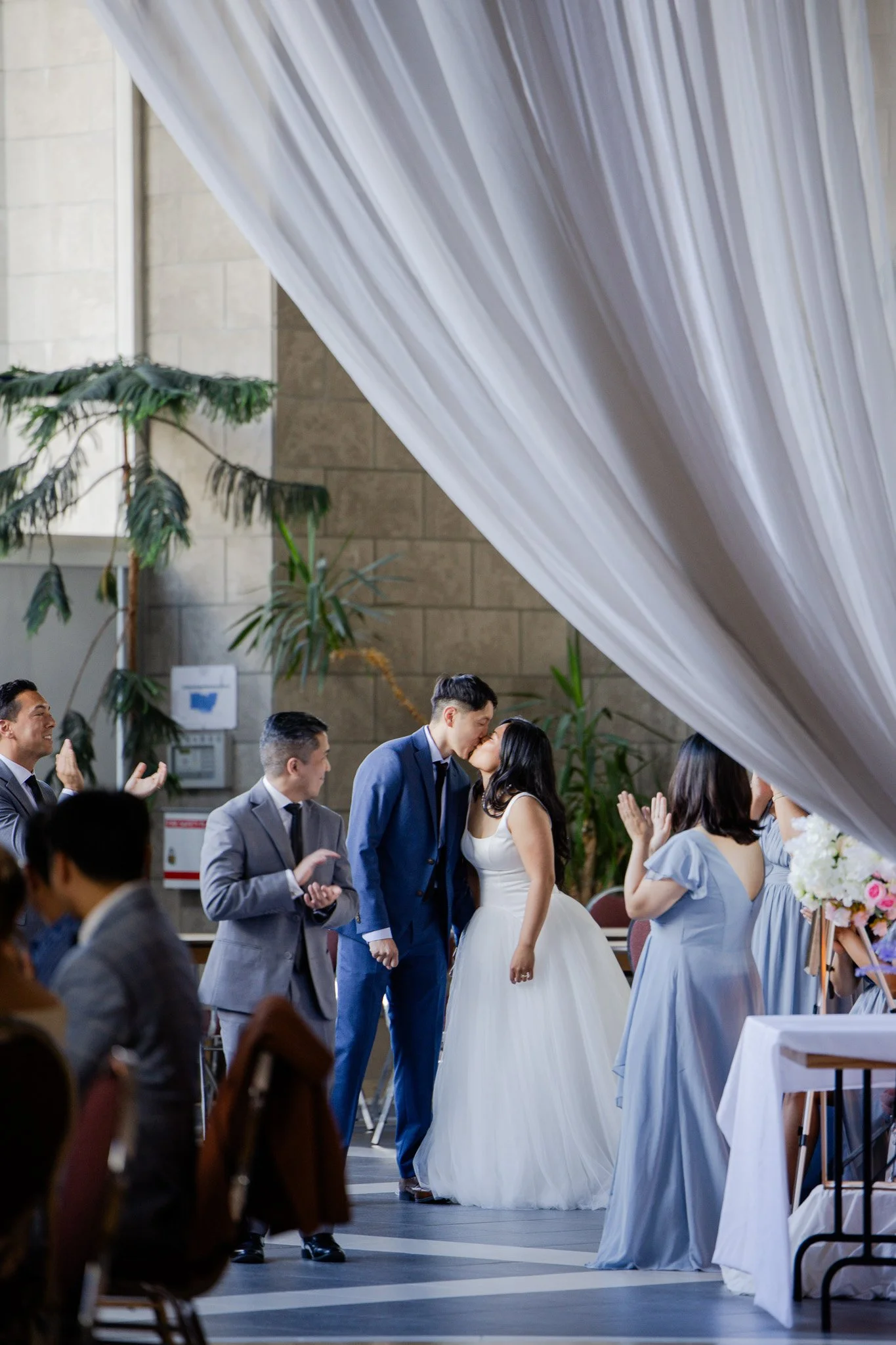 A bride and groom sharing a kiss at their wedding ceremony, surrounded by wedding party members dressed in suits and dresses, with guests seated and clapping in the background.