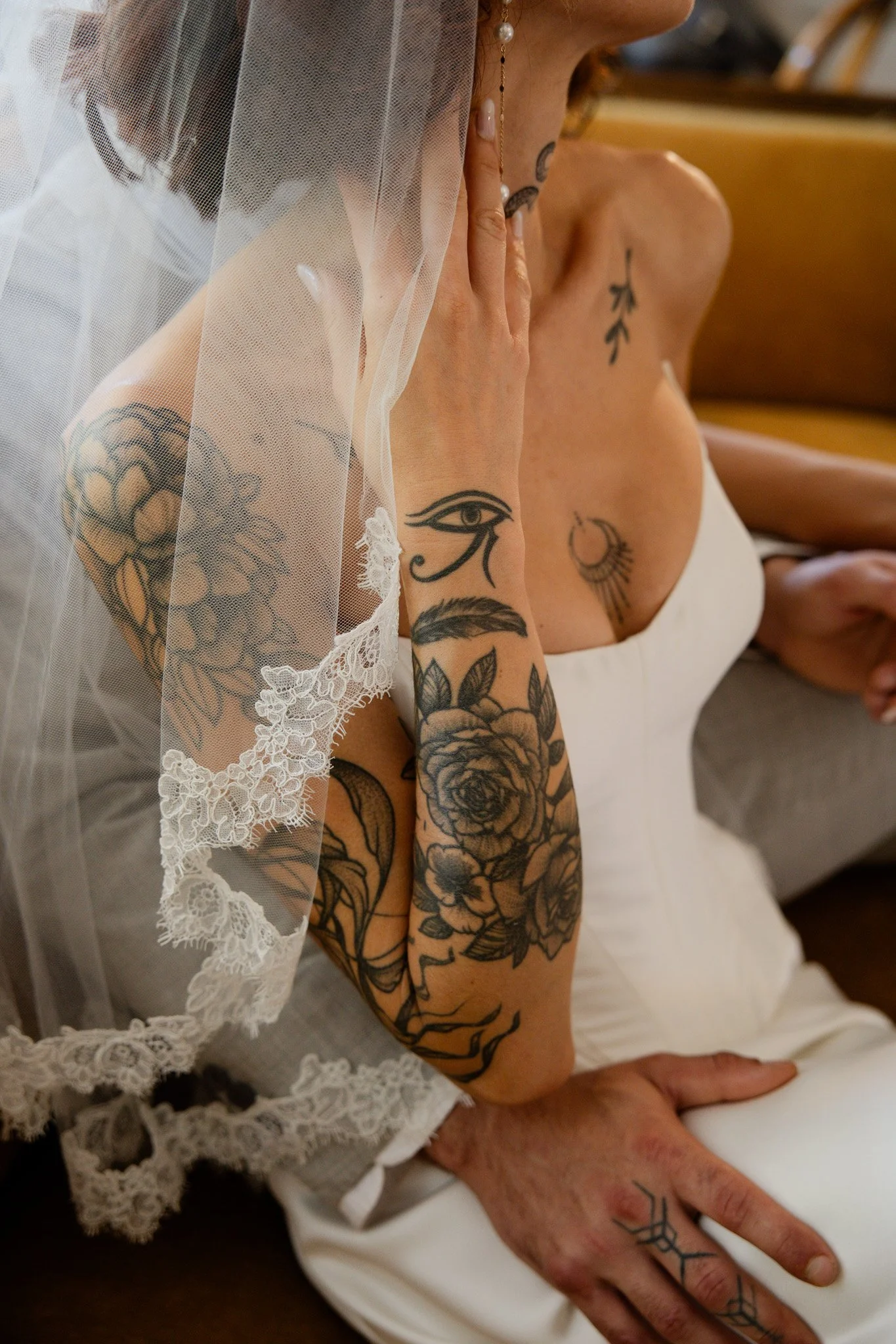 Close-up of a bride with tattoos touching her face, wearing lace trim and a veil, sitting next to a groom with tattoos on his hand and arm, both dressed in white.