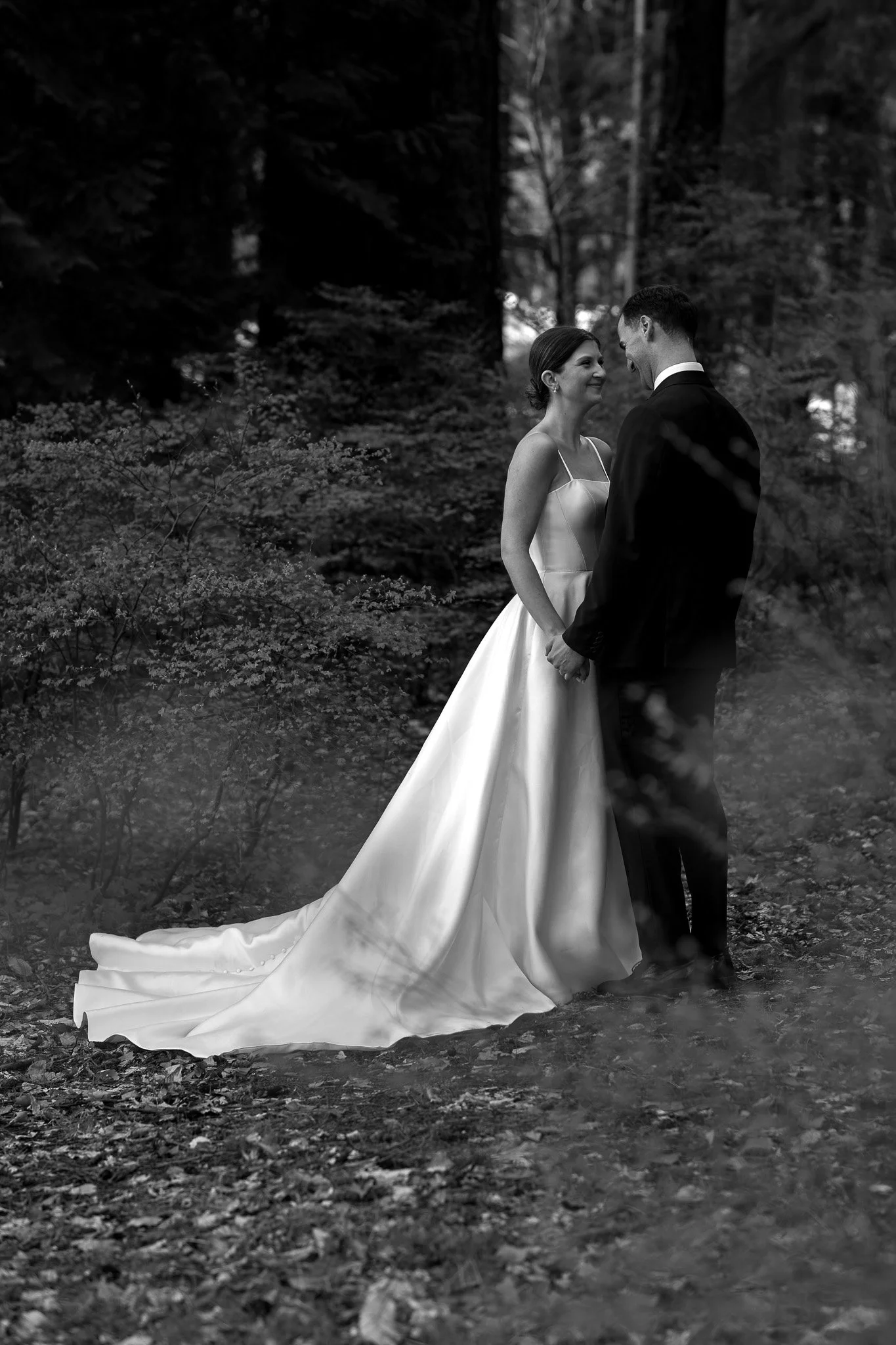 A black and white photo of a bride and groom standing close together outdoors, with trees and foliage in the background. The bride is wearing a wedding dress with a long train, and the groom is in a dark suit. They are smiling and holding hands.