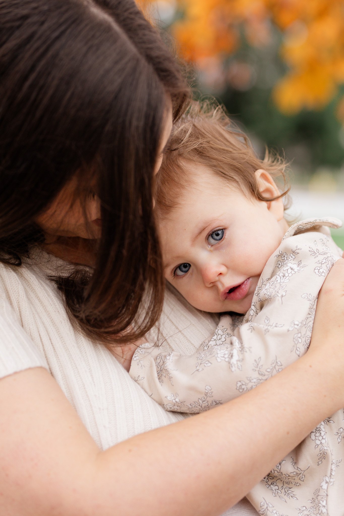 A young woman holding a small child with light brown hair and blue eyes, outdoors during fall with orange leaves in the background.