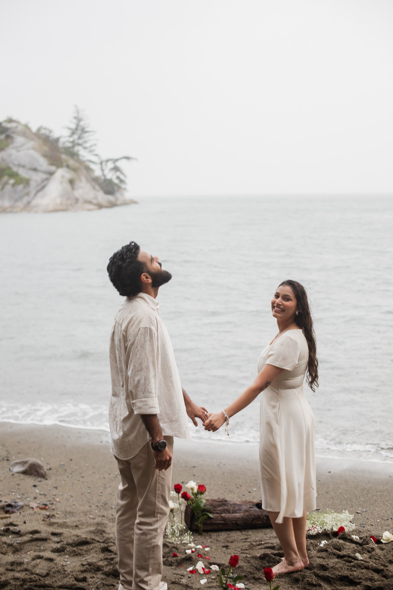 A couple holding hands and smiling at each other on a beach with a rocky outcrop and trees in the background.
