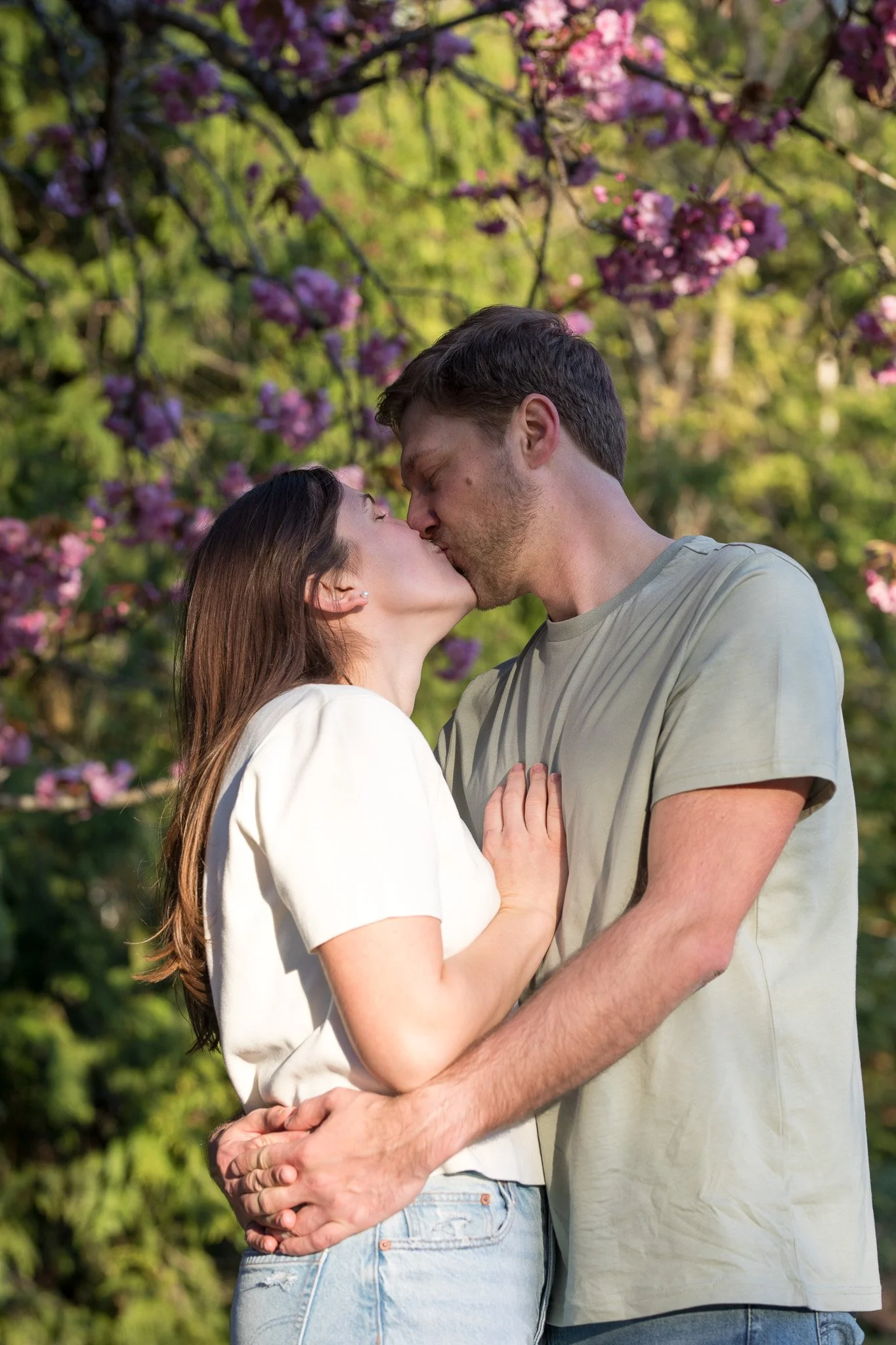 A couple sharing a kiss outdoors under pink blossoms on trees.
