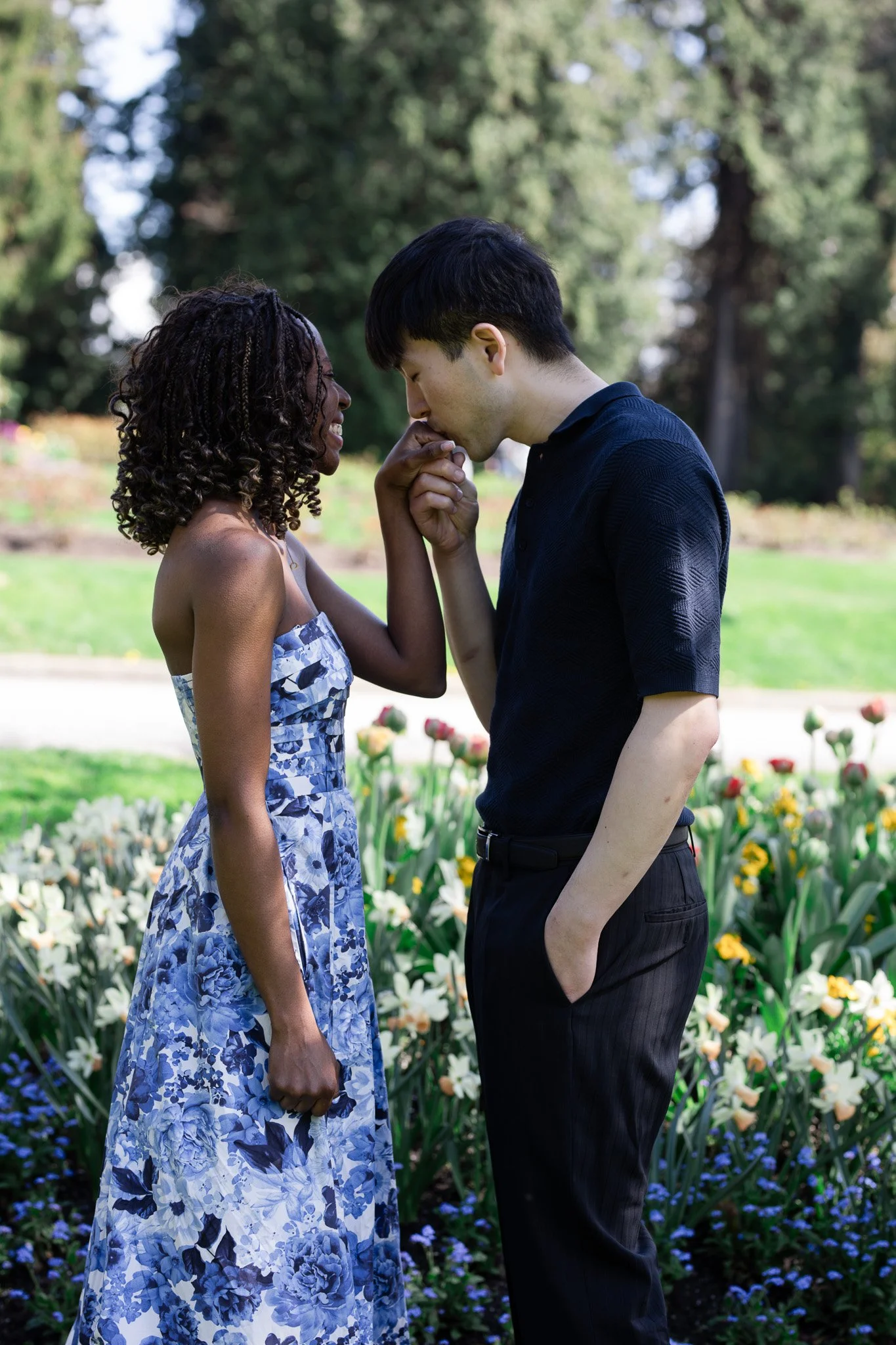 A couple standing in a garden with colorful flowers, with the woman gently kissing the man's hand.