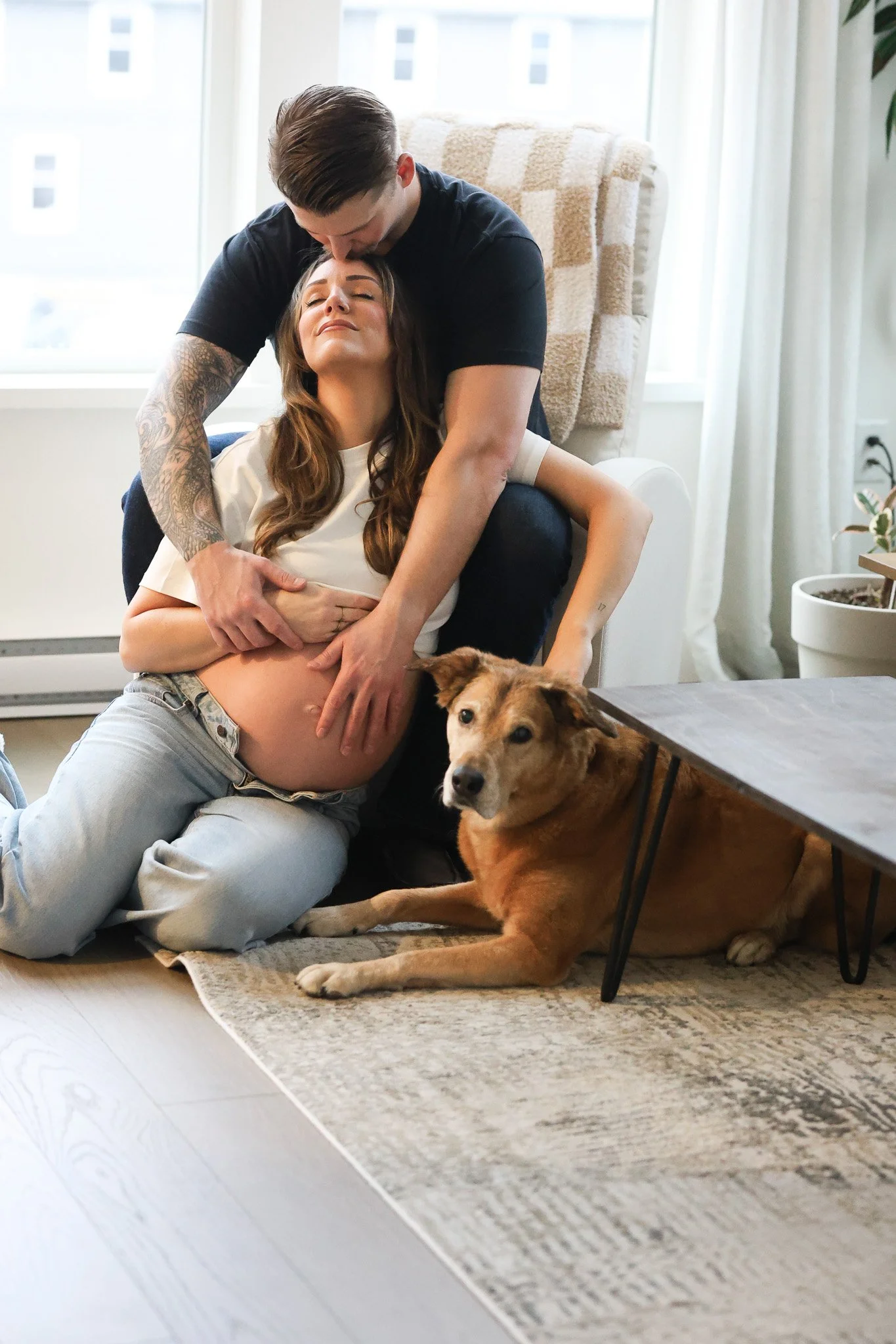 A pregnant woman sitting on the floor with her eyes closed, being embraced from behind by a man who is kissing her forehead. Next to her, a brown dog lies on the rug, looking at the camera. A large window and houseplants are in the background.