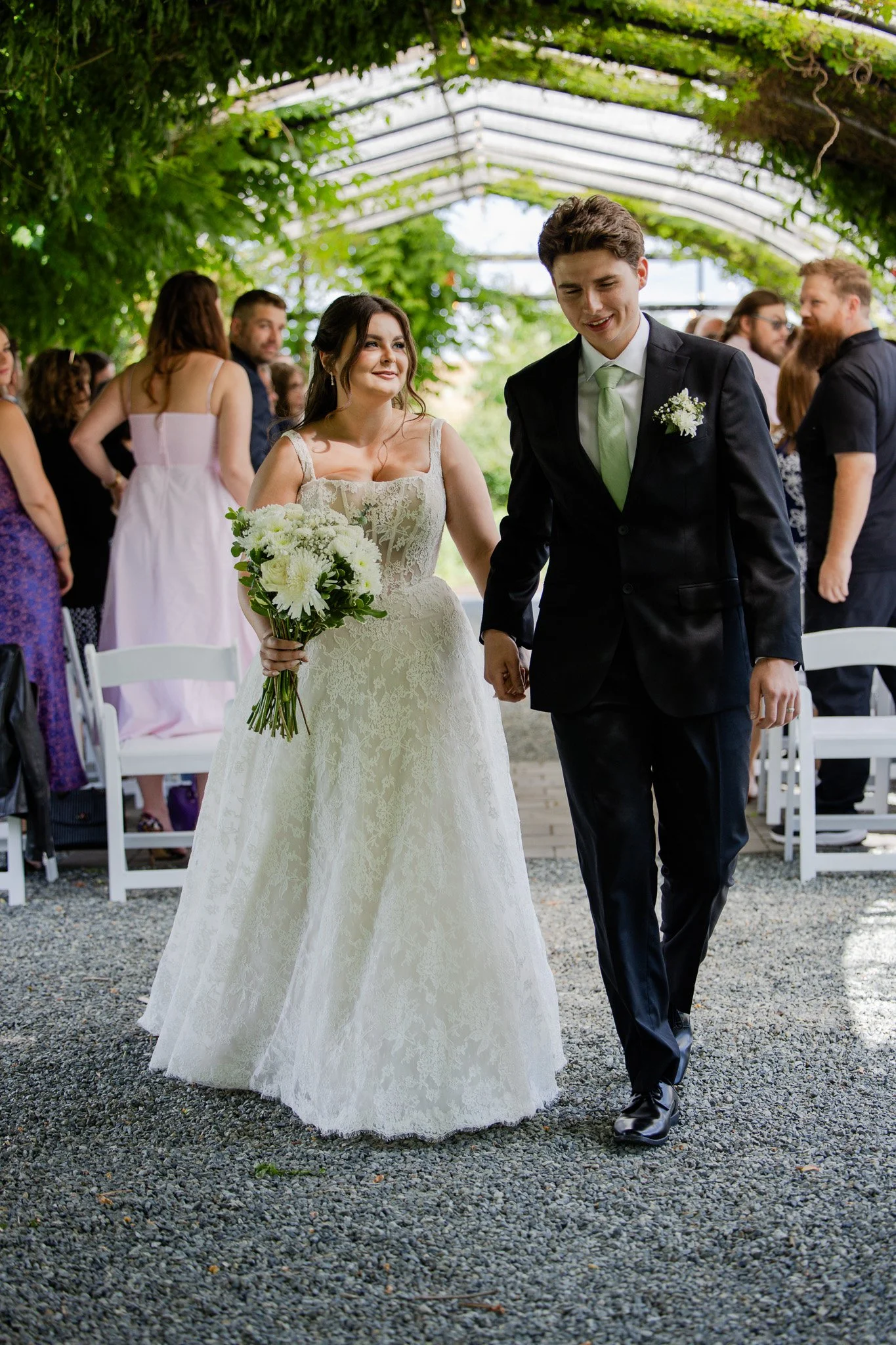 A bride and groom walking hand in hand at their wedding ceremony. The bride is wearing a white lace wedding gown and holding a bouquet of white flowers, while the groom is dressed in a black suit with a light green tie and white boutonniere. Guests are seated and standing under a green, leafy canopy.