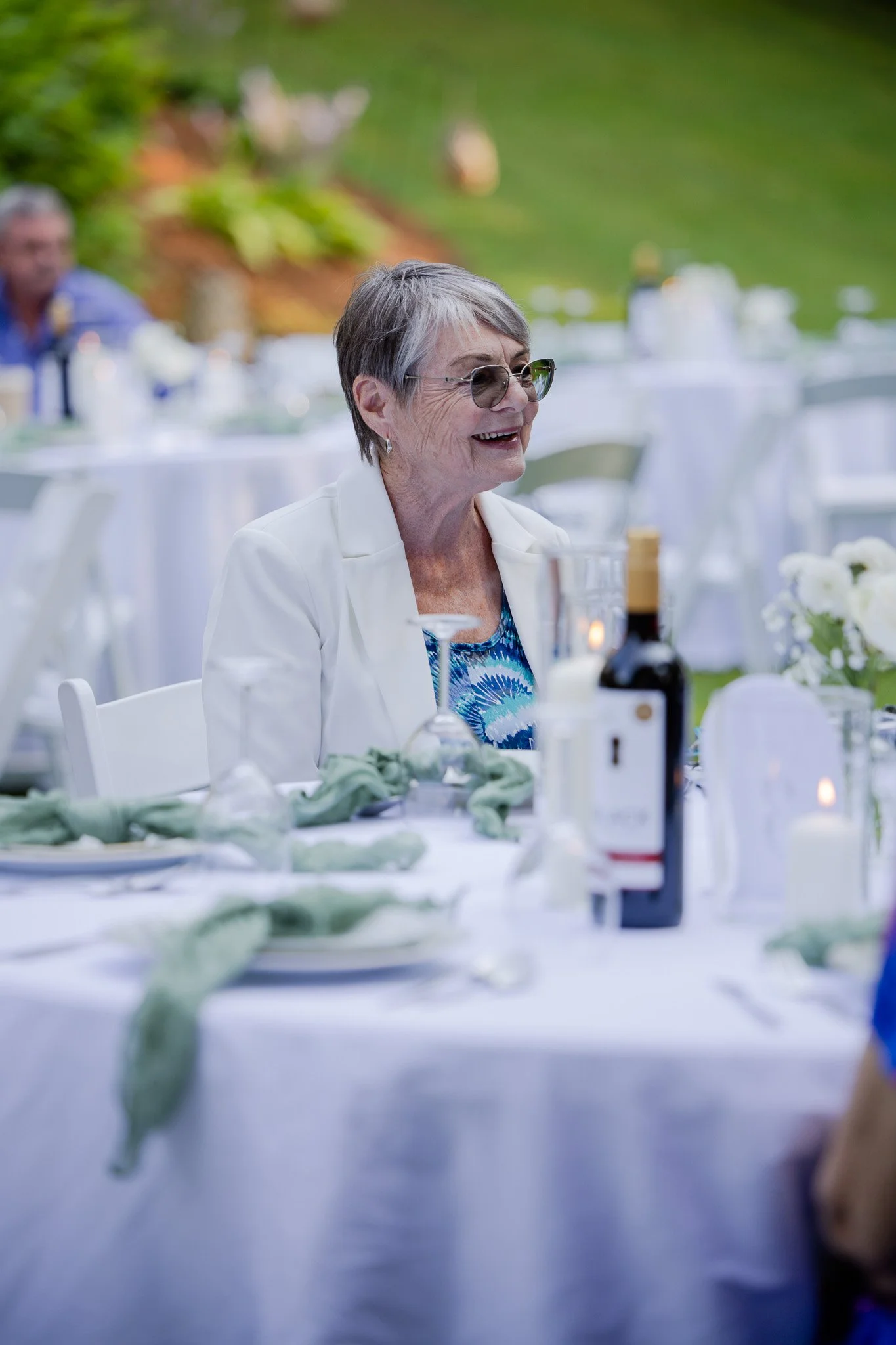 Smiling elderly woman wearing glasses at an outdoor celebration with decorated tables and green background.