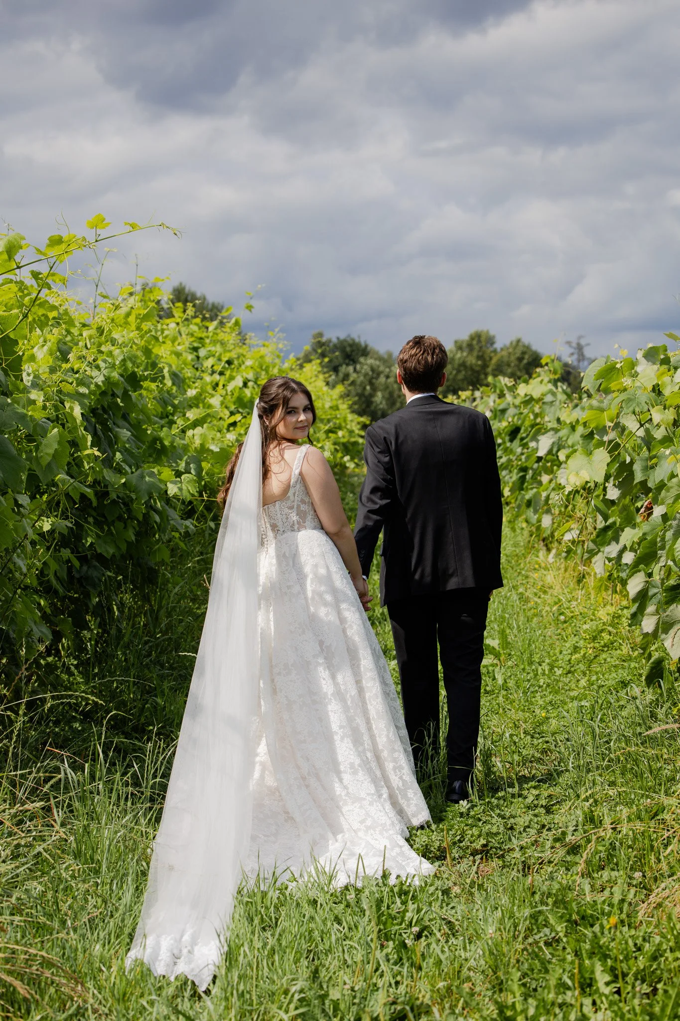 A bride and groom walking hand in hand through a green vineyard under a cloudy sky, with the bride looking back at the camera.